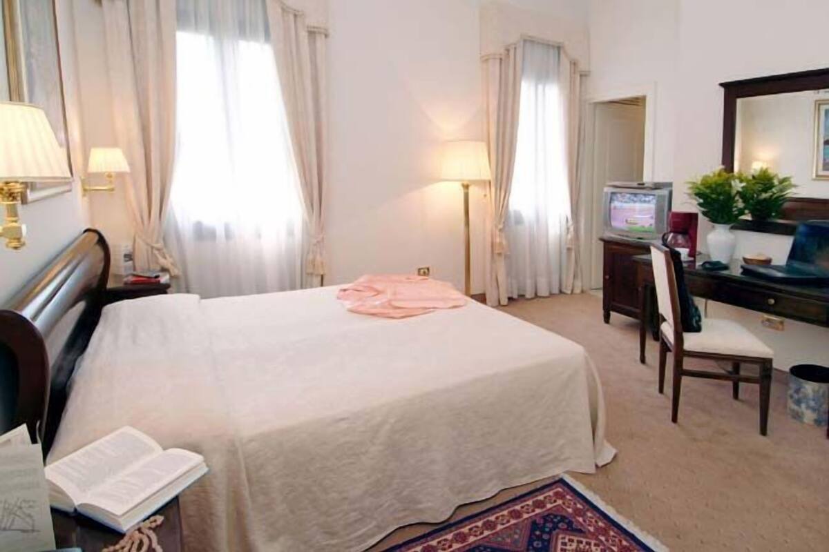 Habitación de hotel con cama, escritorio con televisor, silla y mesita de noche con lámpara.