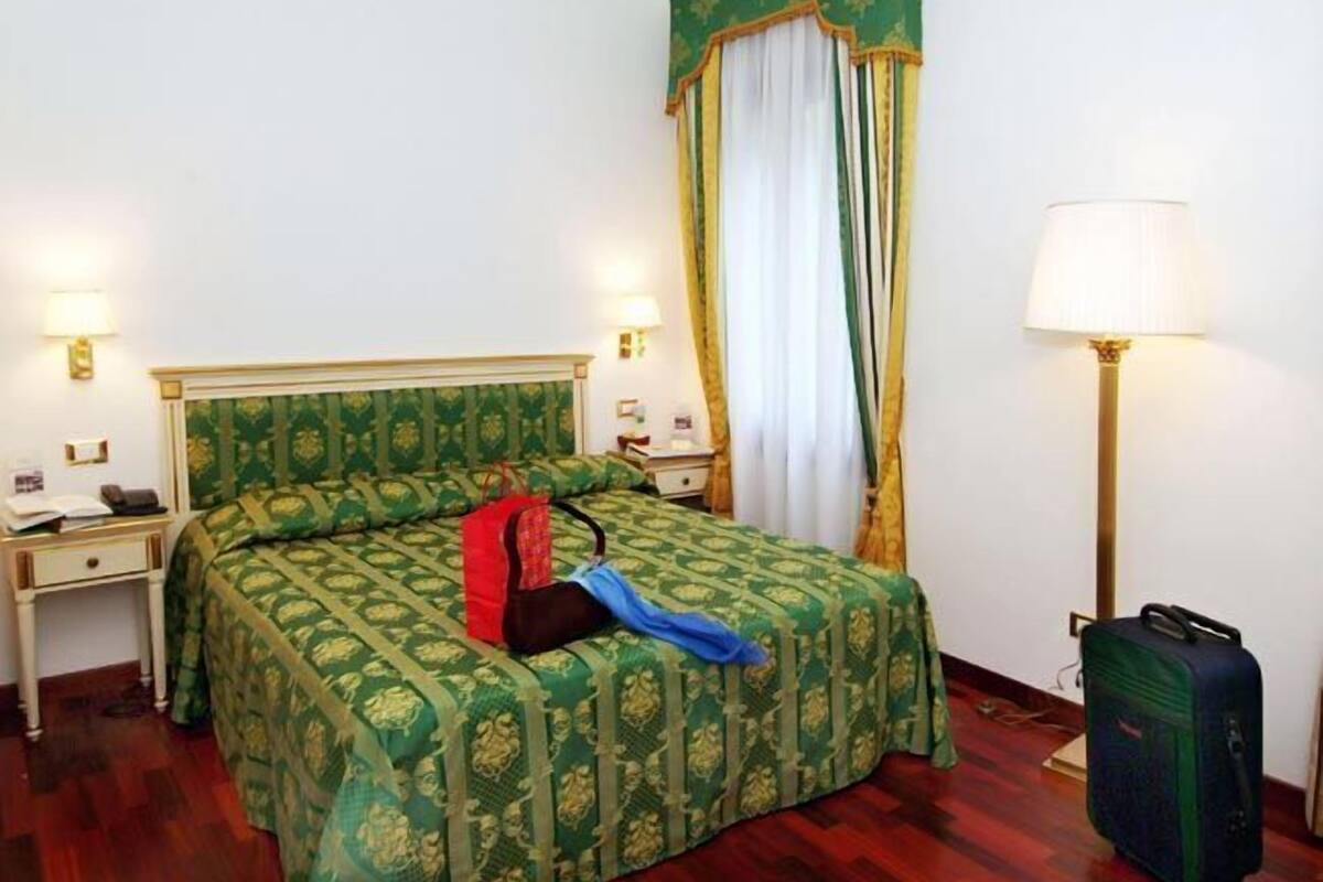 Una habitación de hotel con una cama, una mesita de noche, una lámpara, una maleta y un ventanal con cortinas.