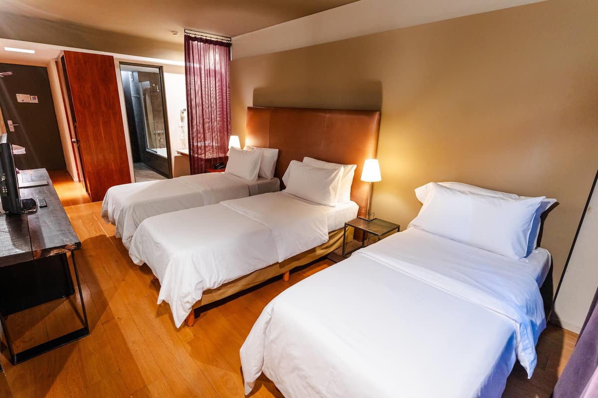 Une chambre d’hôtel avec trois lits simples, un plancher en bois, une tête de lit en bois, un bureau avec une chaise et une salle de bain avec une douche en verre.