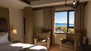 Habitación de hotel con una cama, dos sillones, una lámpara y vistas a un lago y montañas.