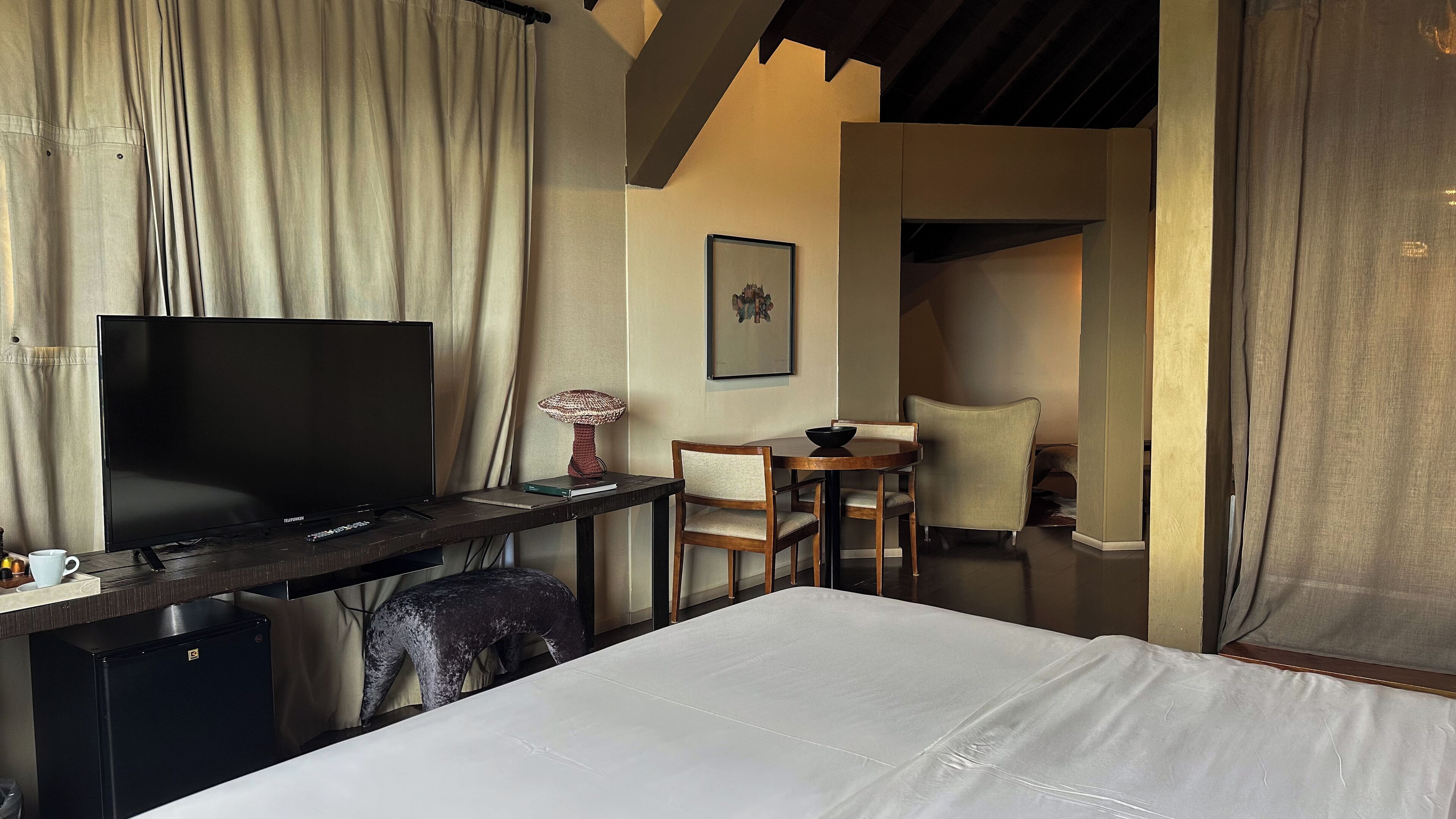 A hotel room with a bed, a desk with a TV, a chair, a small table, and a lamp.