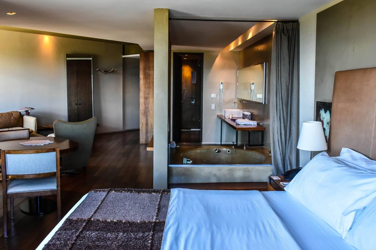 Une chambre d’hôtel dotée d’un grand lit, d’un coin salon avec un fauteuil et une table, d’une salle de bain avec baignoire et d’un bureau avec une lampe.