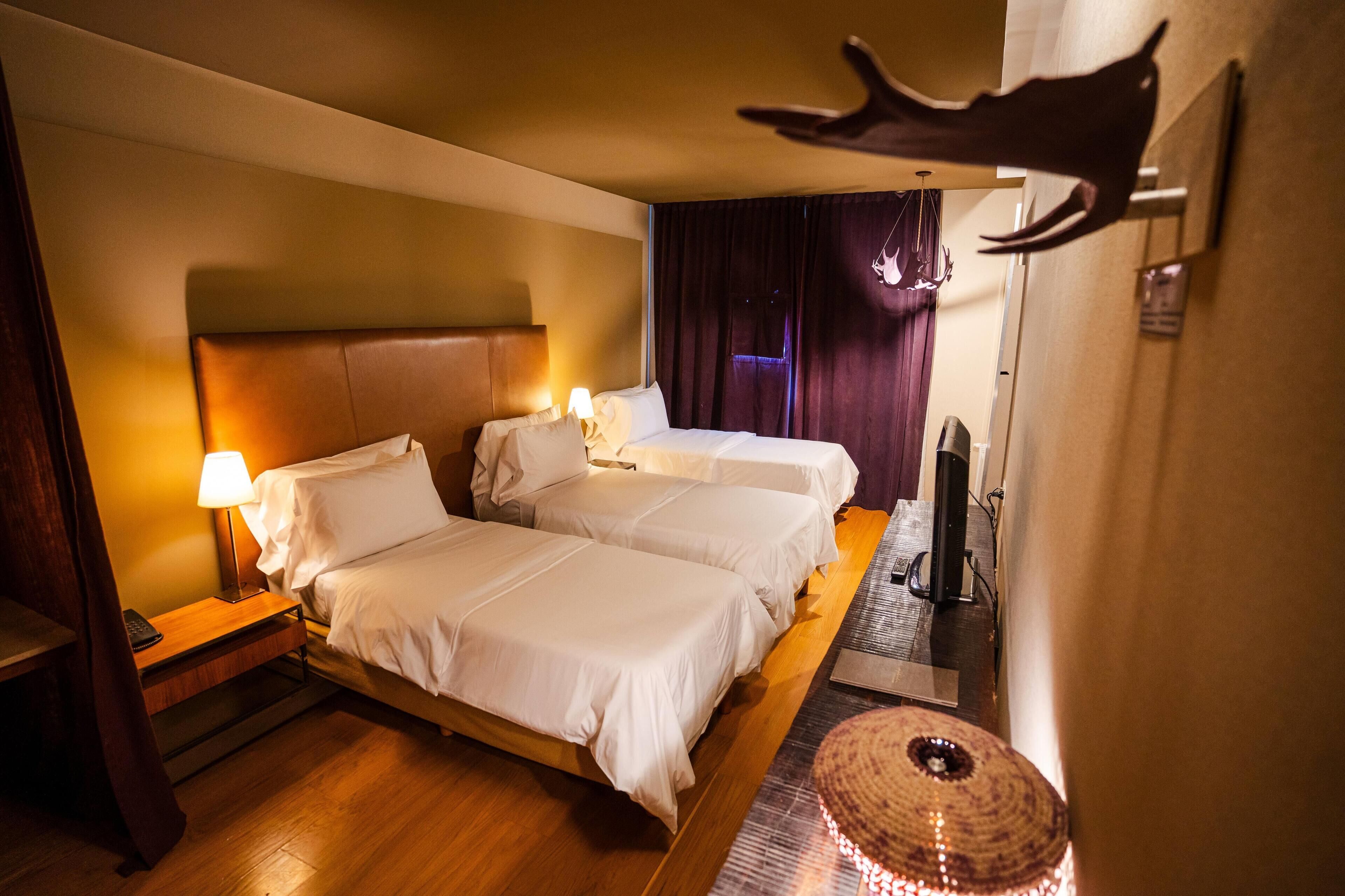 Quarto de hotel com duas camas, uma escrivaninha, uma luminária, uma TV e um objeto decorativo na parede.