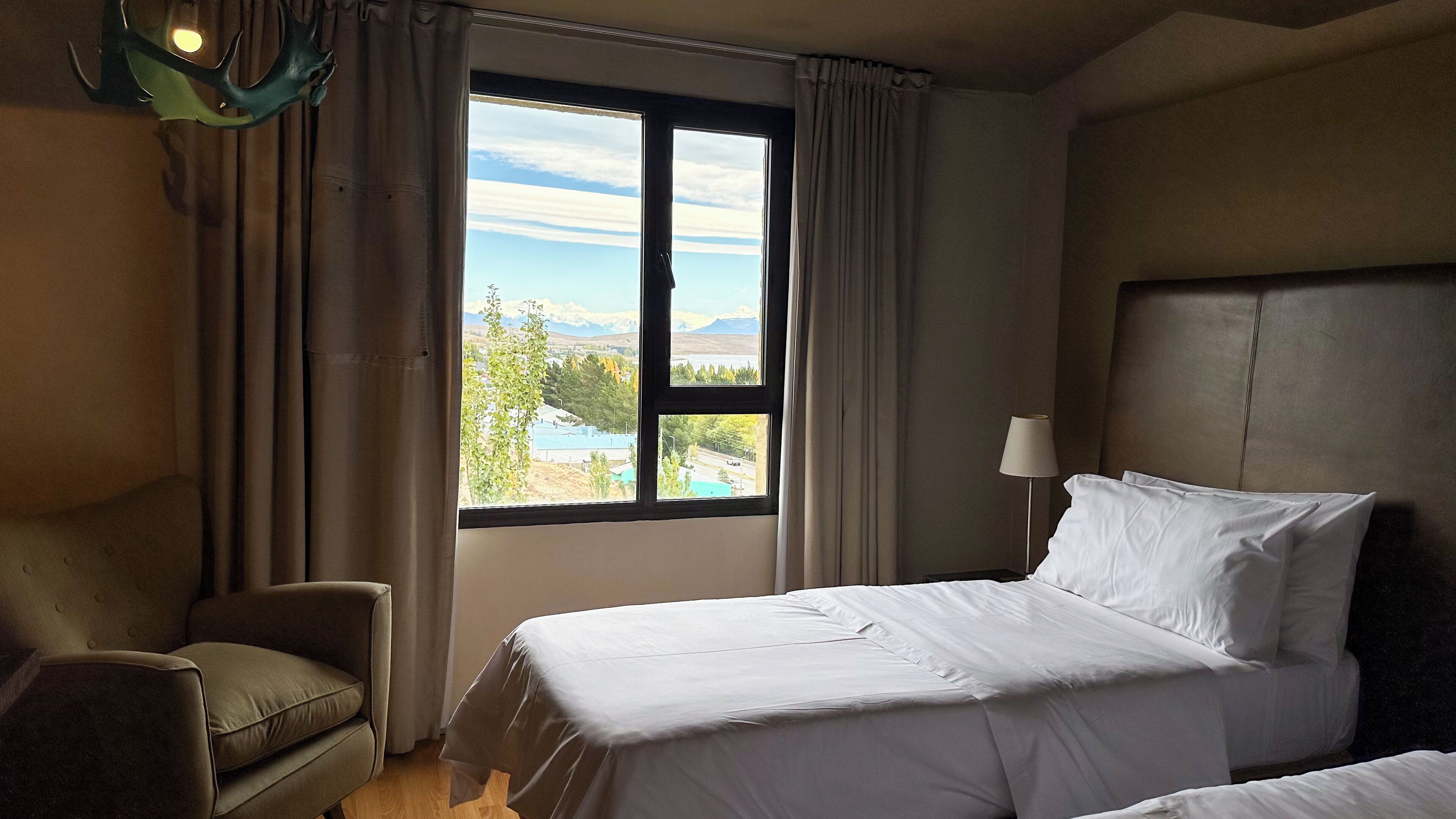 Habitación de hotel con cama, una silla, ventana con vistas a montañas y una piscina, y una lámpara.