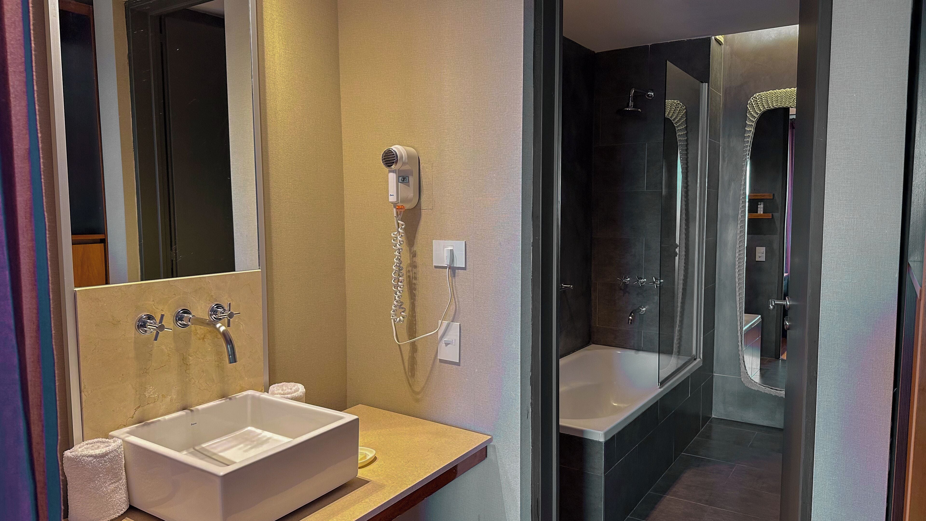A modern bathroom with a large mirror, a sink, a hairdryer, and a glass-enclosed shower.
