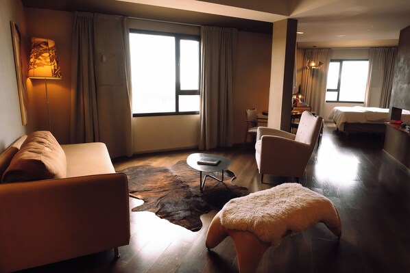 Et moderne hotelværelse med sofa, lænestol, skammel og seng. Der er et lille rundt bord og en lampe. Værelset har store vinduer med gardiner.