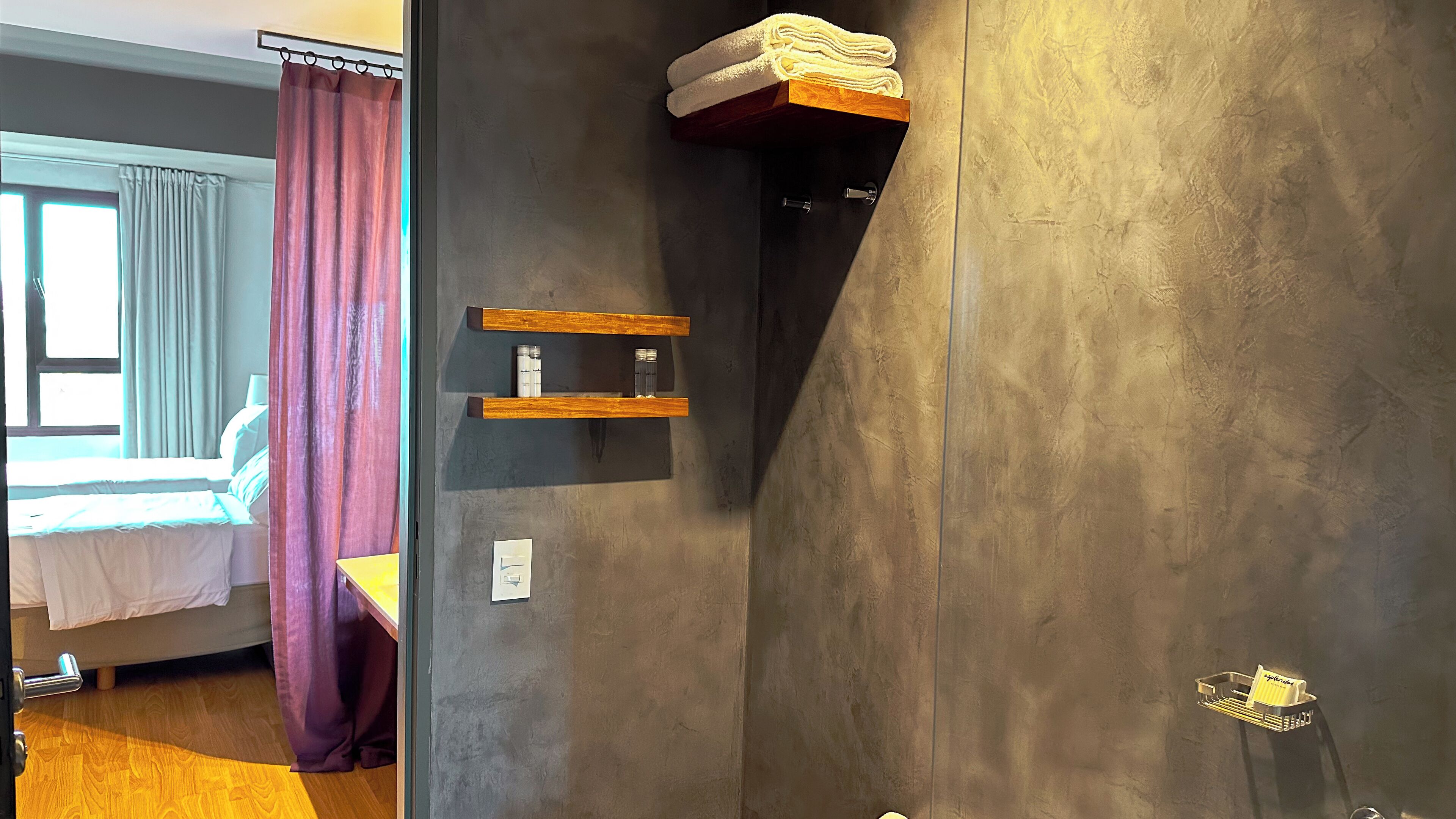 A bathroom with a grey textured wall, a wooden shelf with towels, a small shelf with toiletries, and a door with a keyhole.