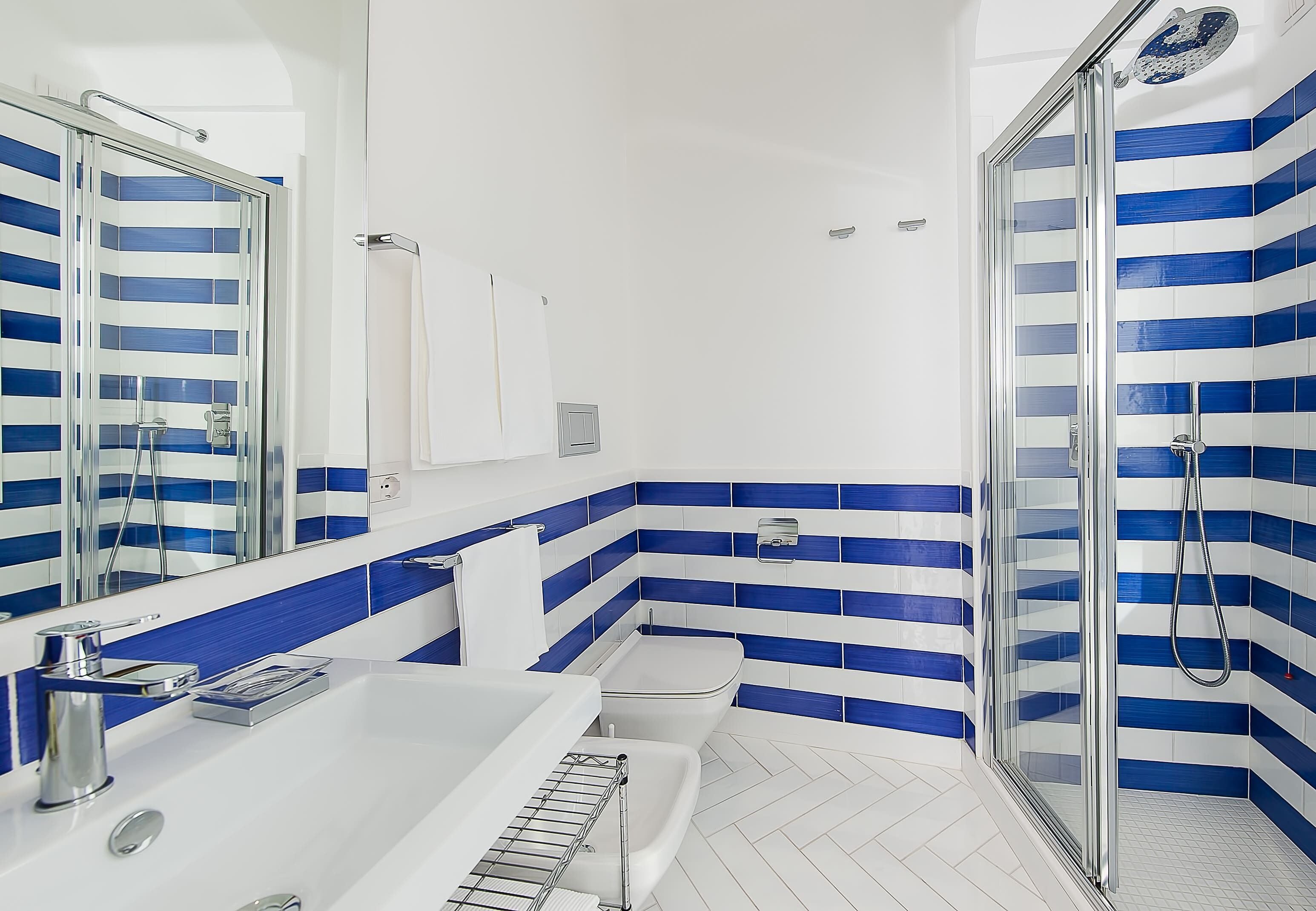 Un baño moderno con un lavamanos blanco, una cabina de ducha con mampara de vidrio y azulejos a rayas azules.