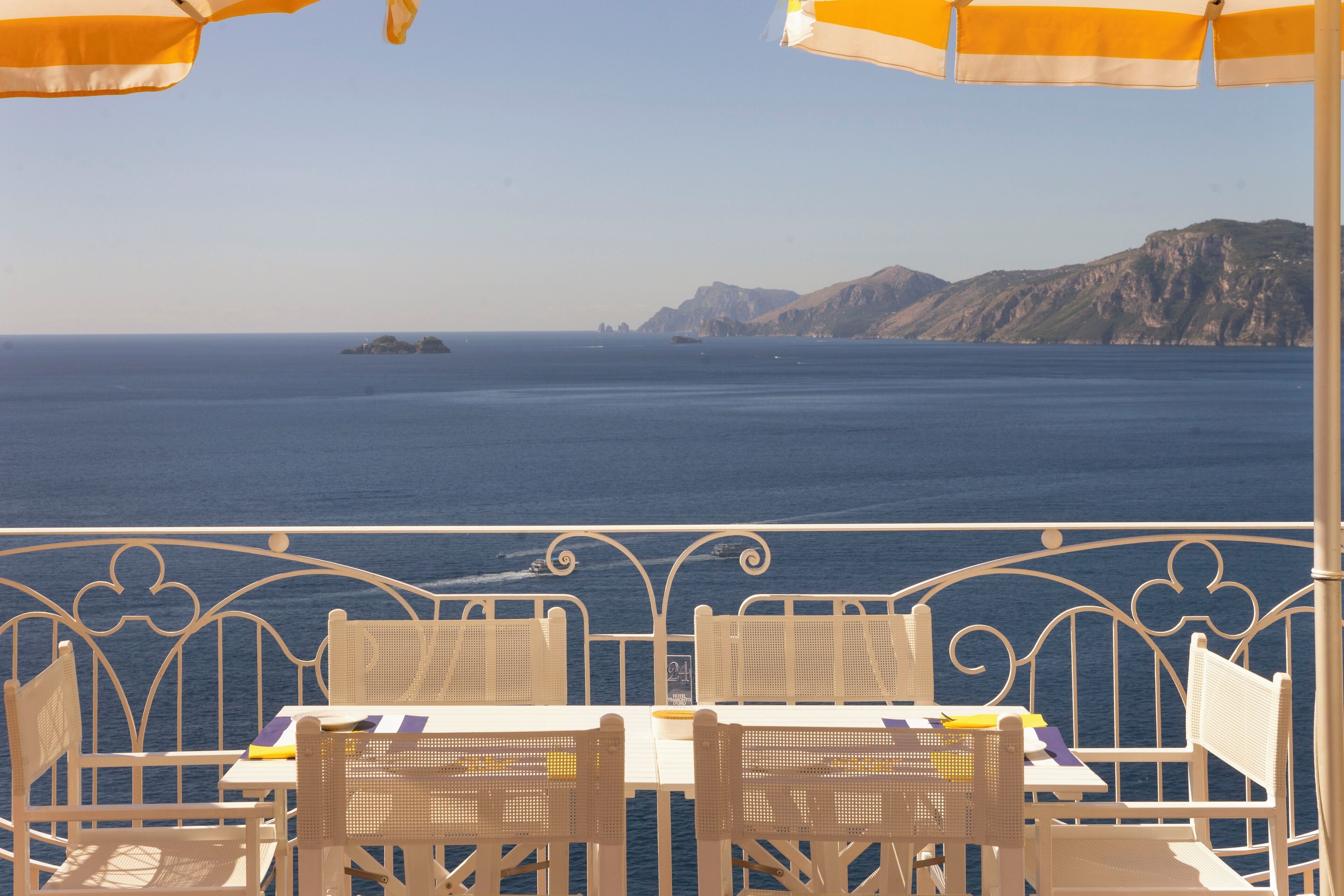 A seaside terrace with white metal chairs and tables, overlooking a calm sea and distant rocky coastline.