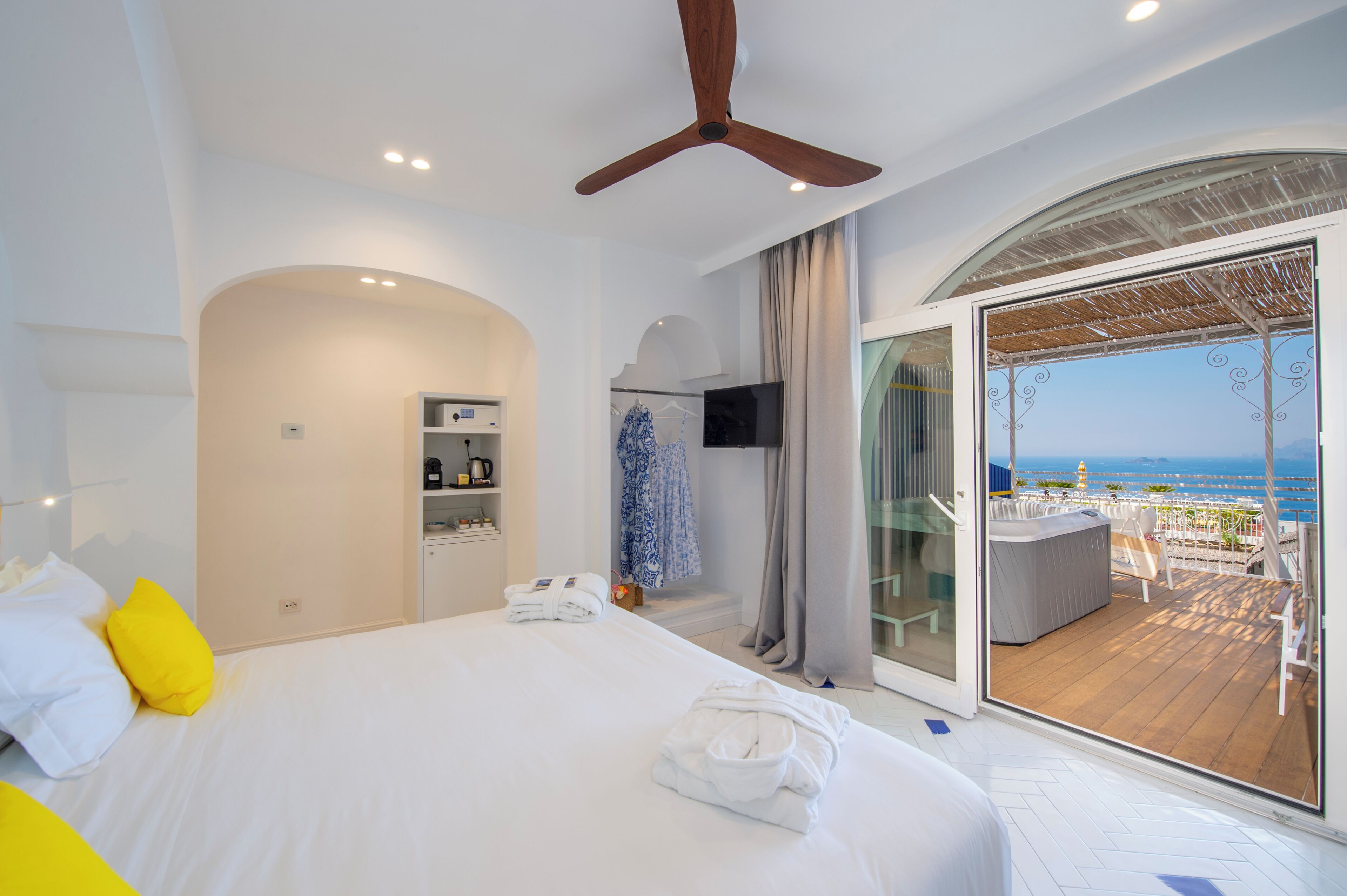 Habitación de hotel con cama, ventilador de techo, televisor, minibar y balcón con vista al mar.