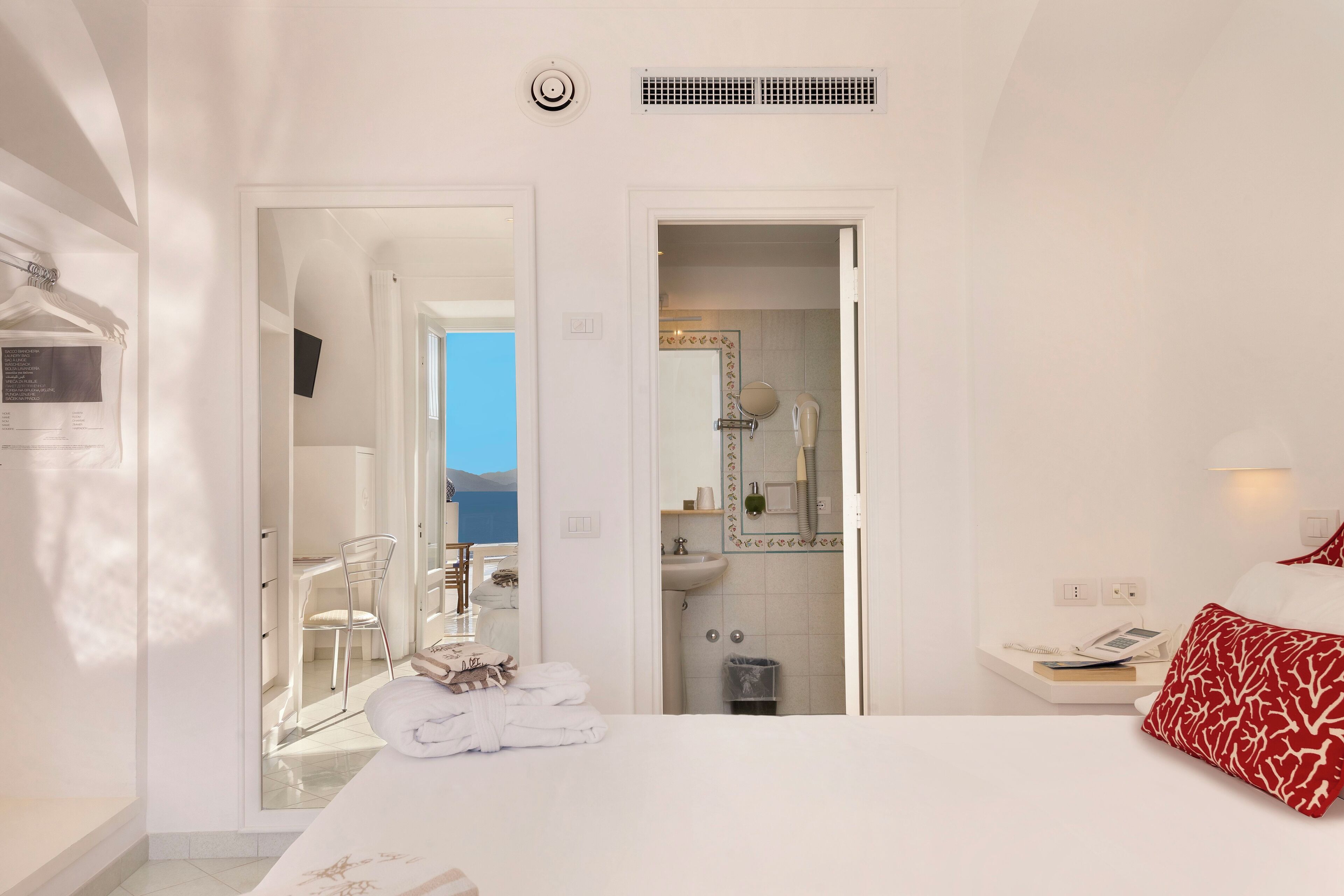 Habitación de hotel con cama, mesita de noche con teléfono y libros, baño con espejo y lavamanos, y vista al cielo.