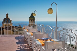 Una terraza con sillas y mesas de metal blanco, vista al mar, e iglesia con cúpula y campanario.