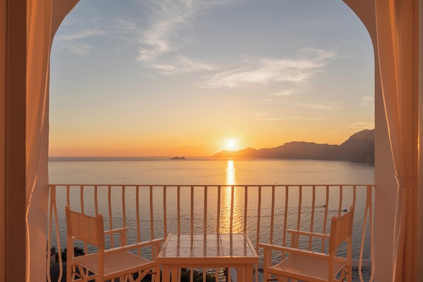 Un balcón con una mesa y dos sillas, con vista a la puesta de sol sobre un mar tranquilo y formas de tierra en la distancia.
