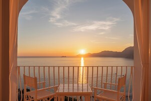 Un balcón con una mesa y dos sillas, con vista a la puesta de sol sobre un mar tranquilo y formas de tierra en la distancia.