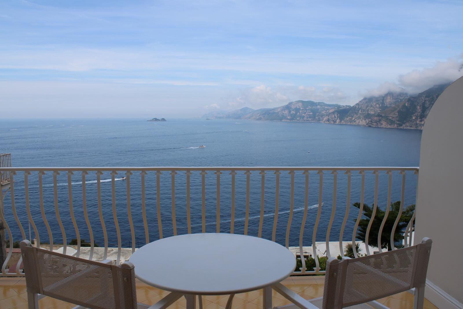 Un balcón con una mesa blanca y dos sillas de mimbre, con vistas a un mar tranquilo con islas y montañas a lo lejos.