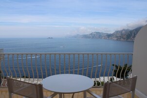 Un balcón con una mesa blanca y dos sillas de mimbre, con vistas a un mar tranquilo con islas y montañas a lo lejos.