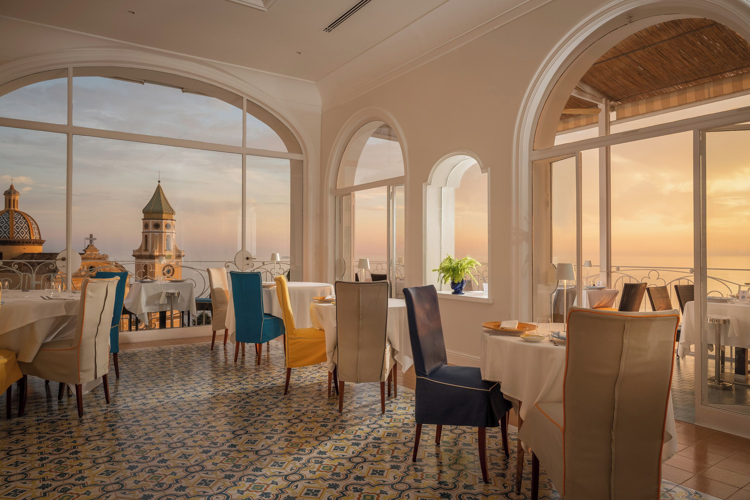 A dining area with tables and chairs, large arched windows, and a patterned floor.