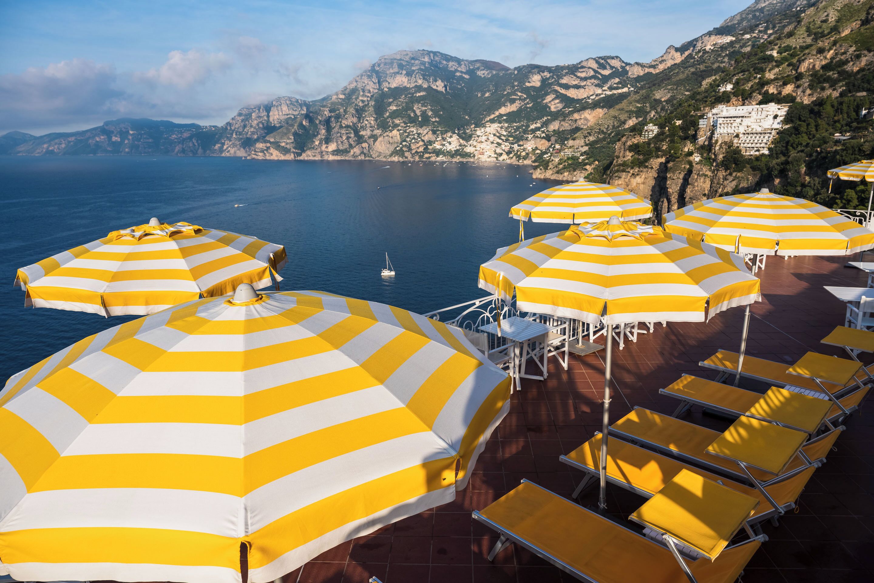 A terrace with yellow and white striped umbrellas and sun loungers overlooking a body of water and mountains.