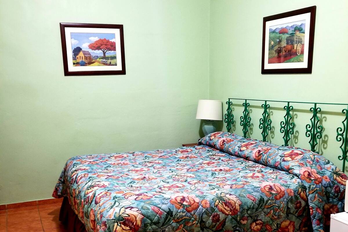A room with a bed, a chair, a table, and two framed pictures on the wall.