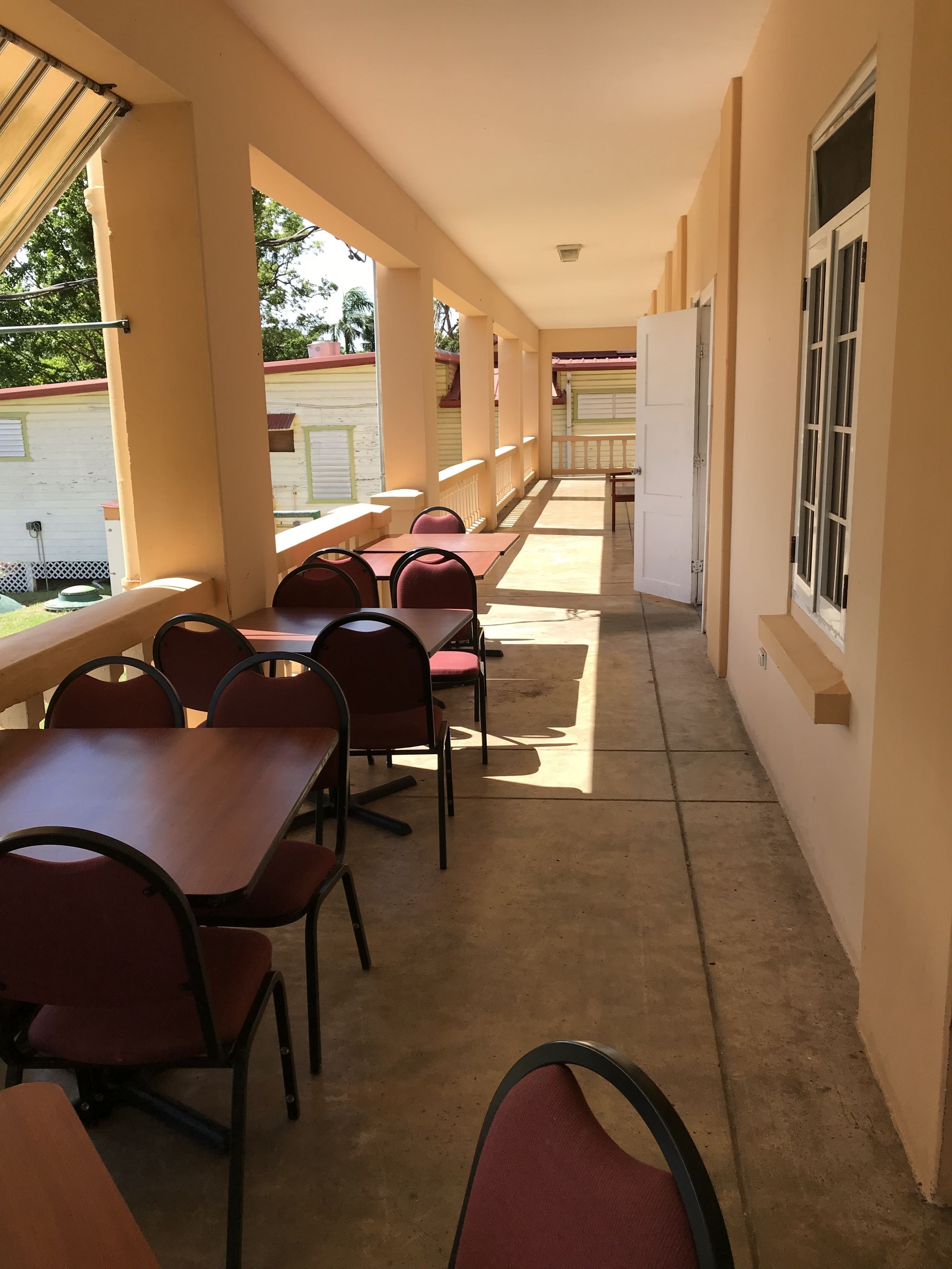 Un área de estar al aire libre techada con mesas y sillas, piso de concreto y vista a un edificio al fondo.