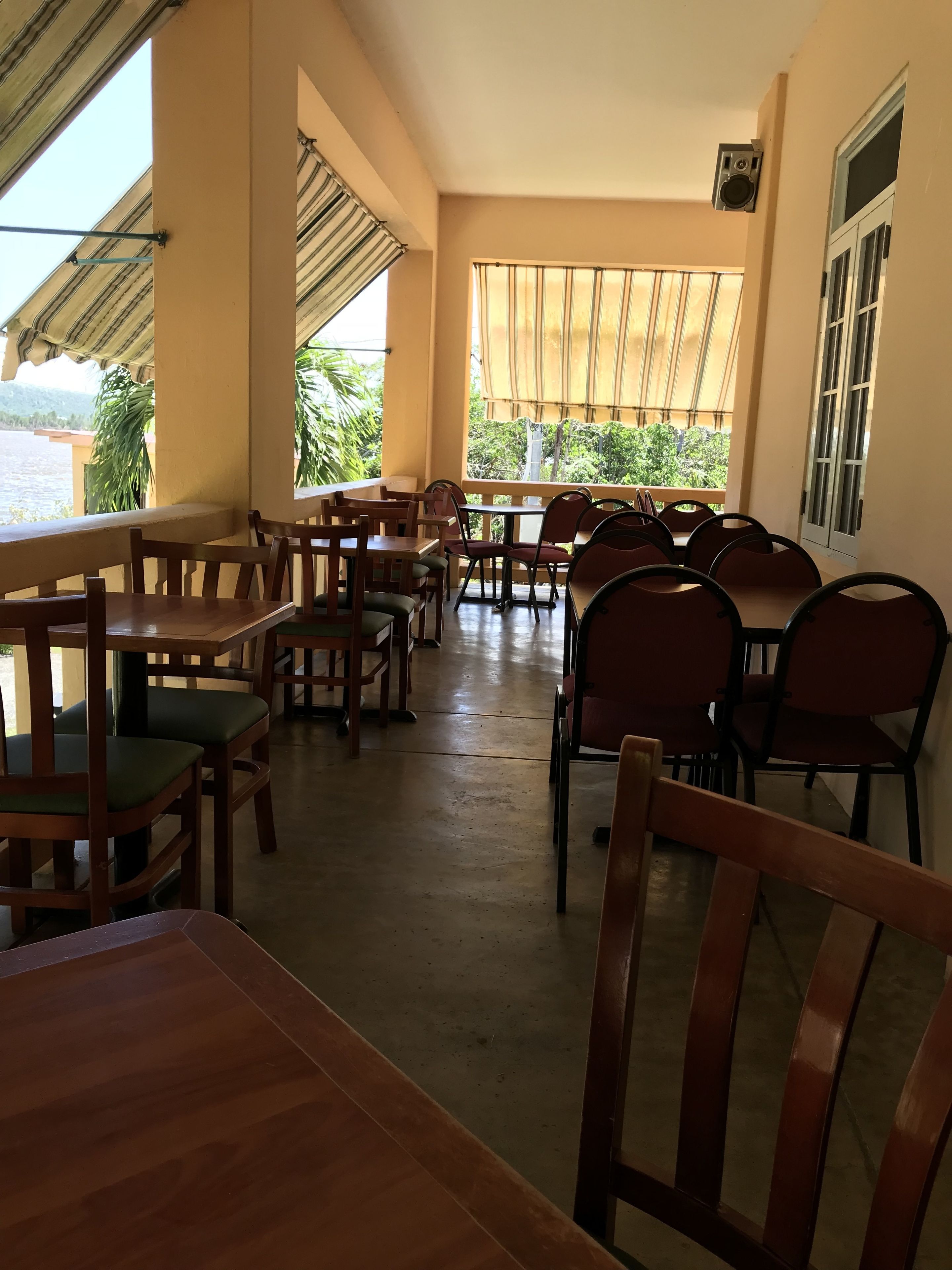 Un restaurant avec des tables et des chaises en bois, de grandes fenêtres ornées de rideaux et une vue sur la verdure et l’eau.