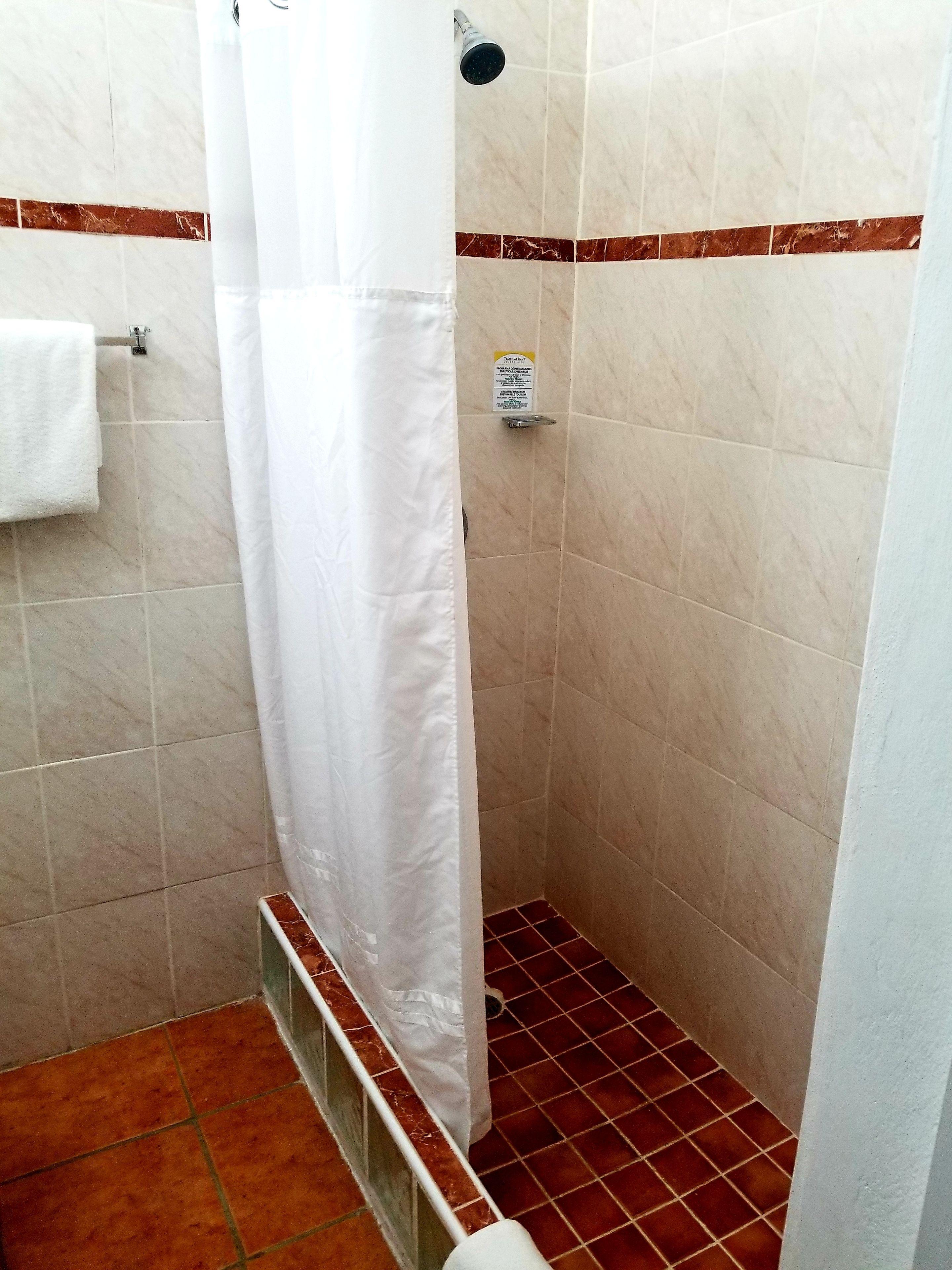 Pequeño espacio de ducha con azulejos, cortina blanca, una toalla colgada en un perchero y un letrero en la pared.