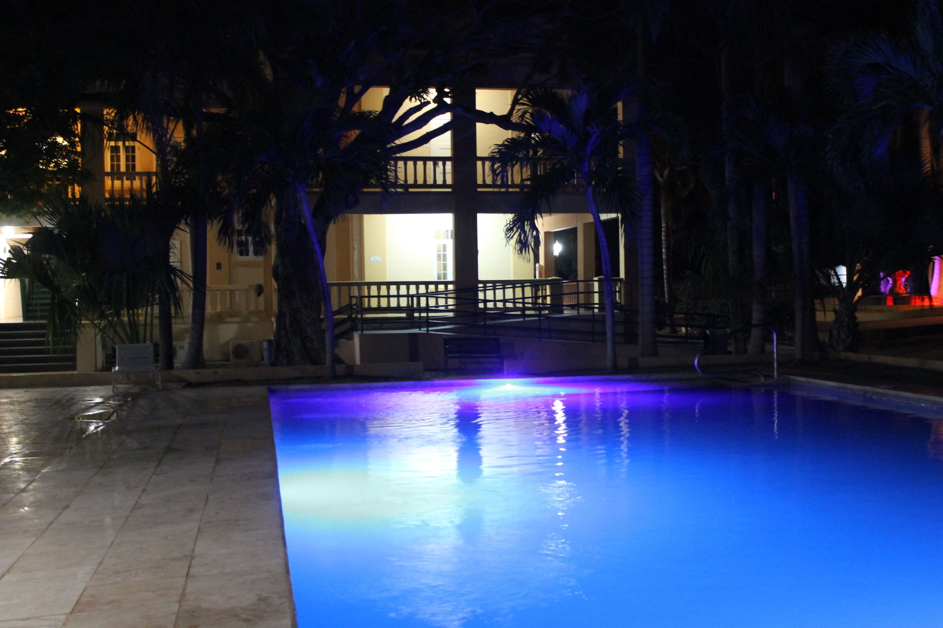 A swimming pool with a blue-lit edge, surrounded by palm trees and a building with a balcony in the background.