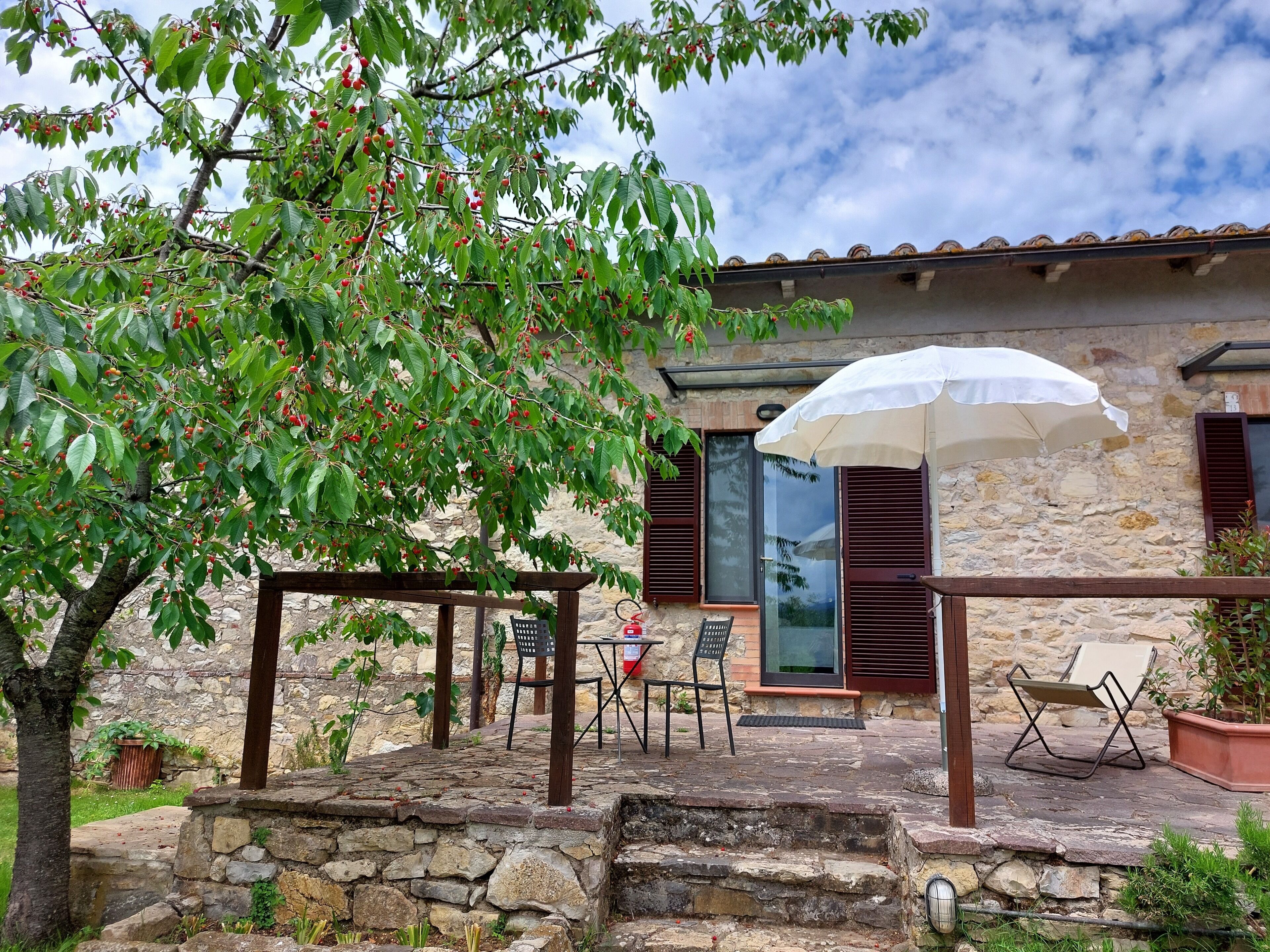 Un edificio de piedra con un patio, mobiliario exterior y un árbol con bayas rojas.