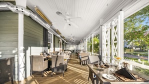 A covered outdoor dining area with wicker furniture and white railings, overlooking a garden and street. - The Shaw Club Hotel (Niagara-on-the-Lake)