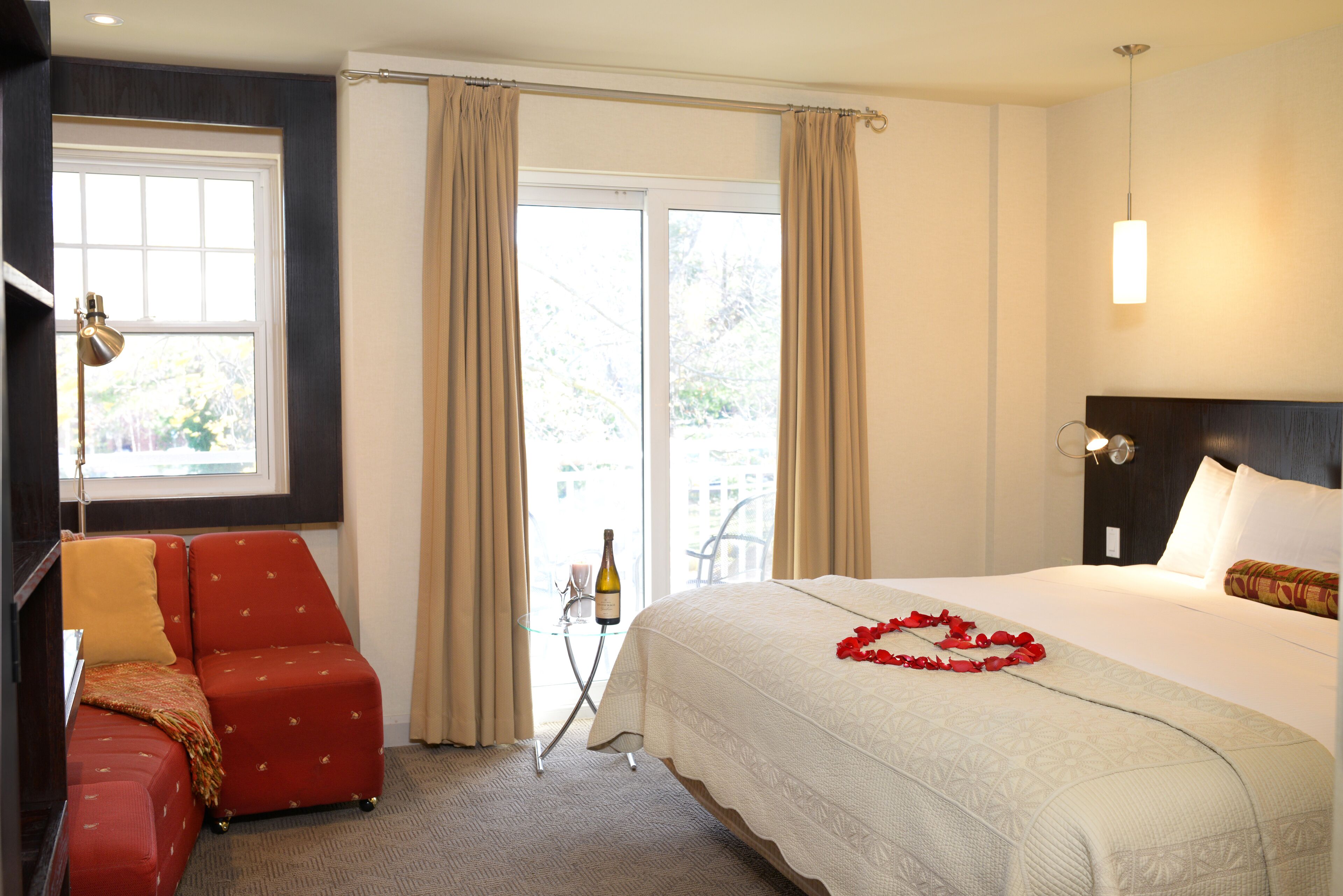 Quarto de hotel com uma cama, uma poltrona vermelha, uma mesinha de centro com uma garrafa e uma vela, e vista pela janela.