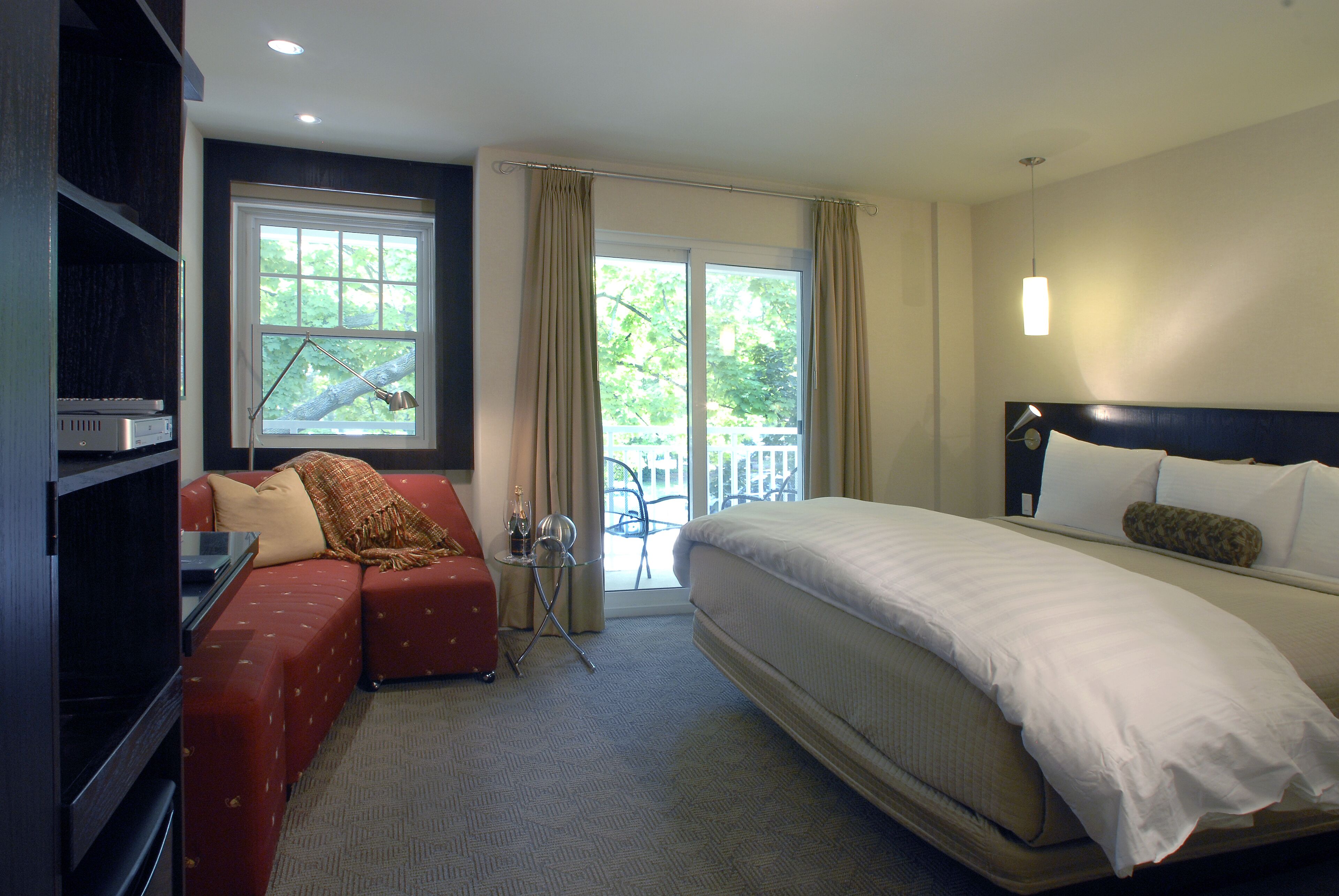 A hotel room with a large bed, a red sofa, a small table with a lamp, and a balcony with a view of trees.