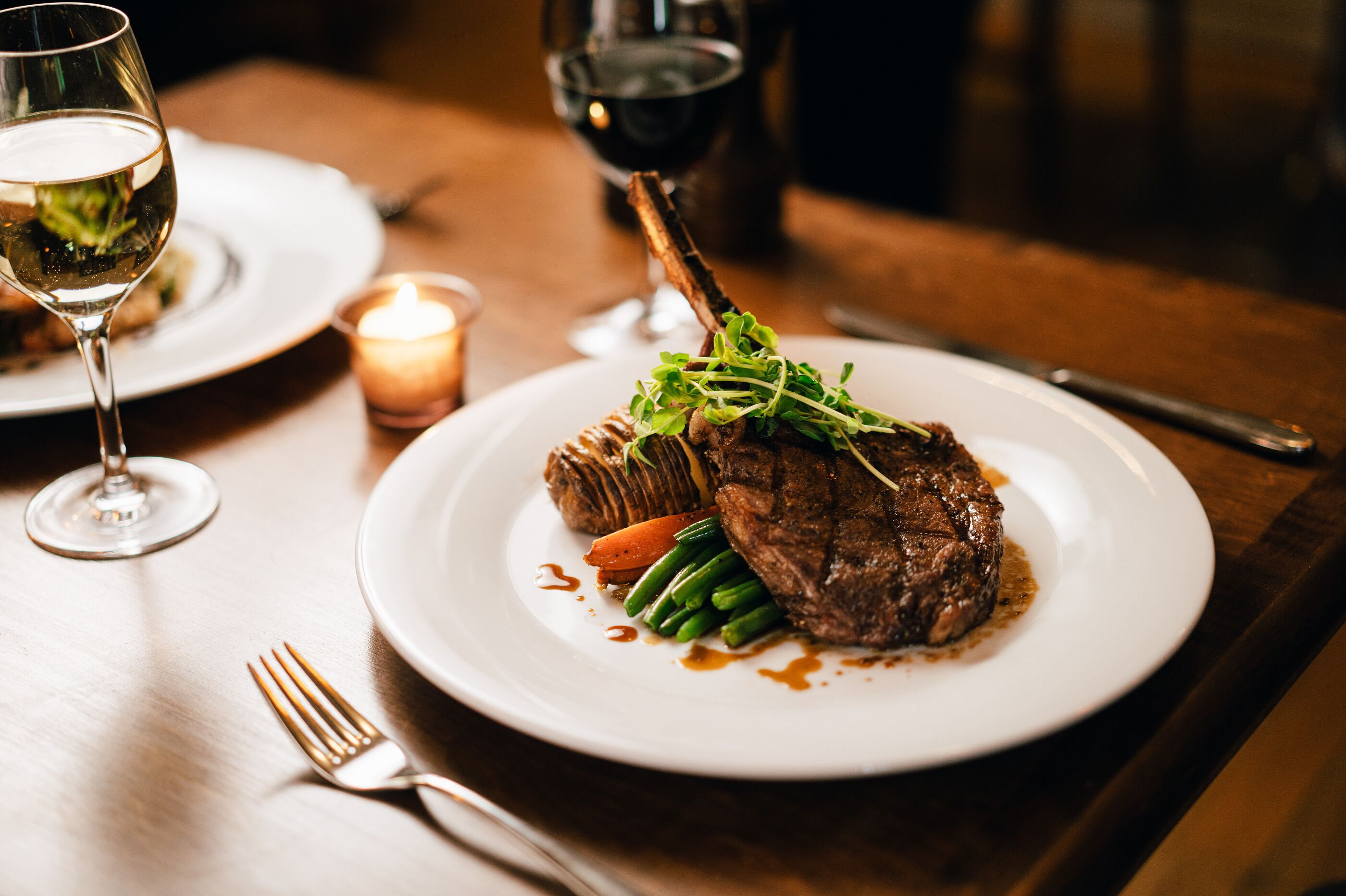 A plate of steak with vegetables and sauce, a glass of white wine, and a candle on a wooden table.