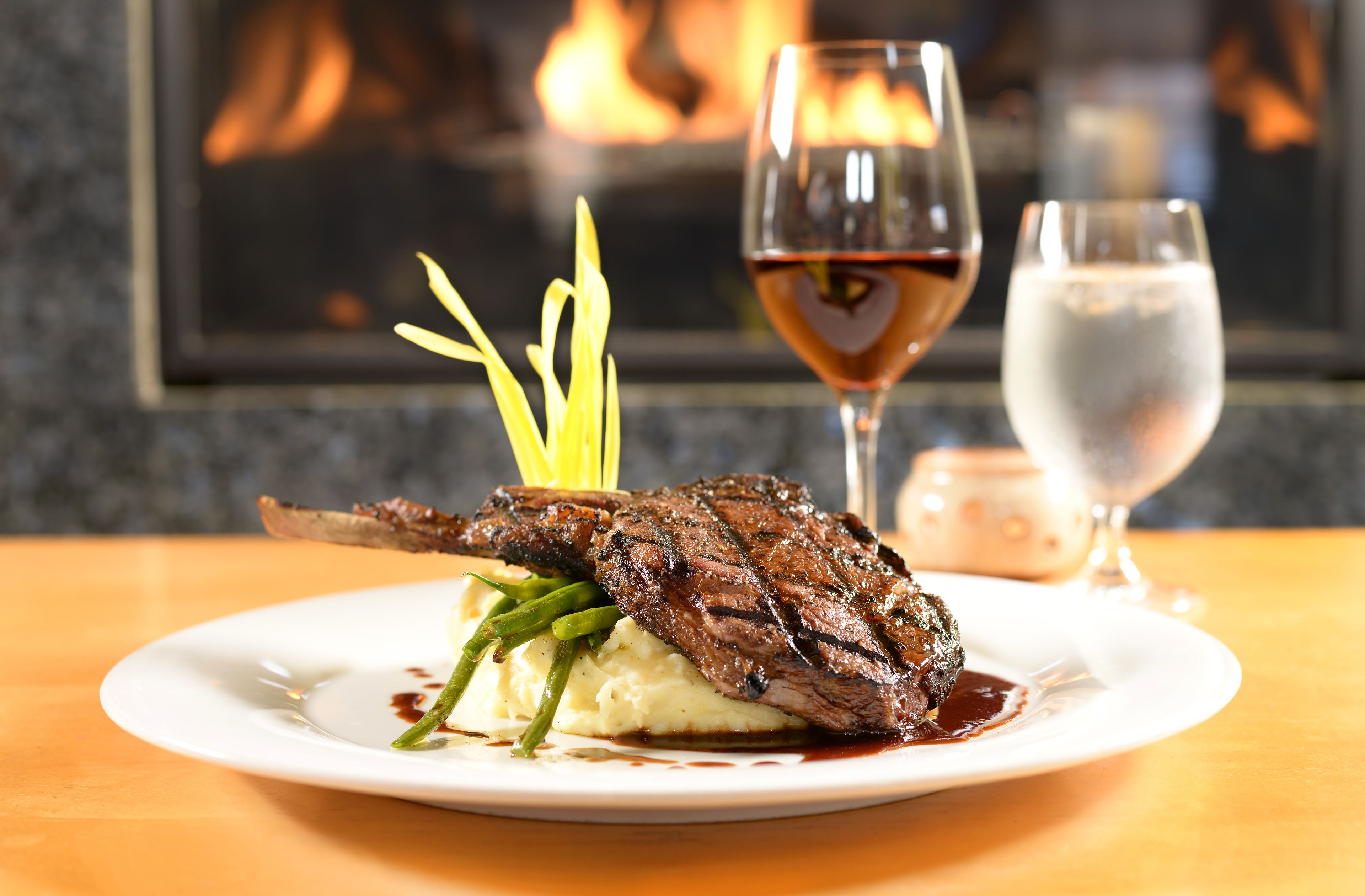 A plate of steak with green beans and mashed potatoes, accompanied by a glass of red wine and a glass of water, set against a background with a fireplace.