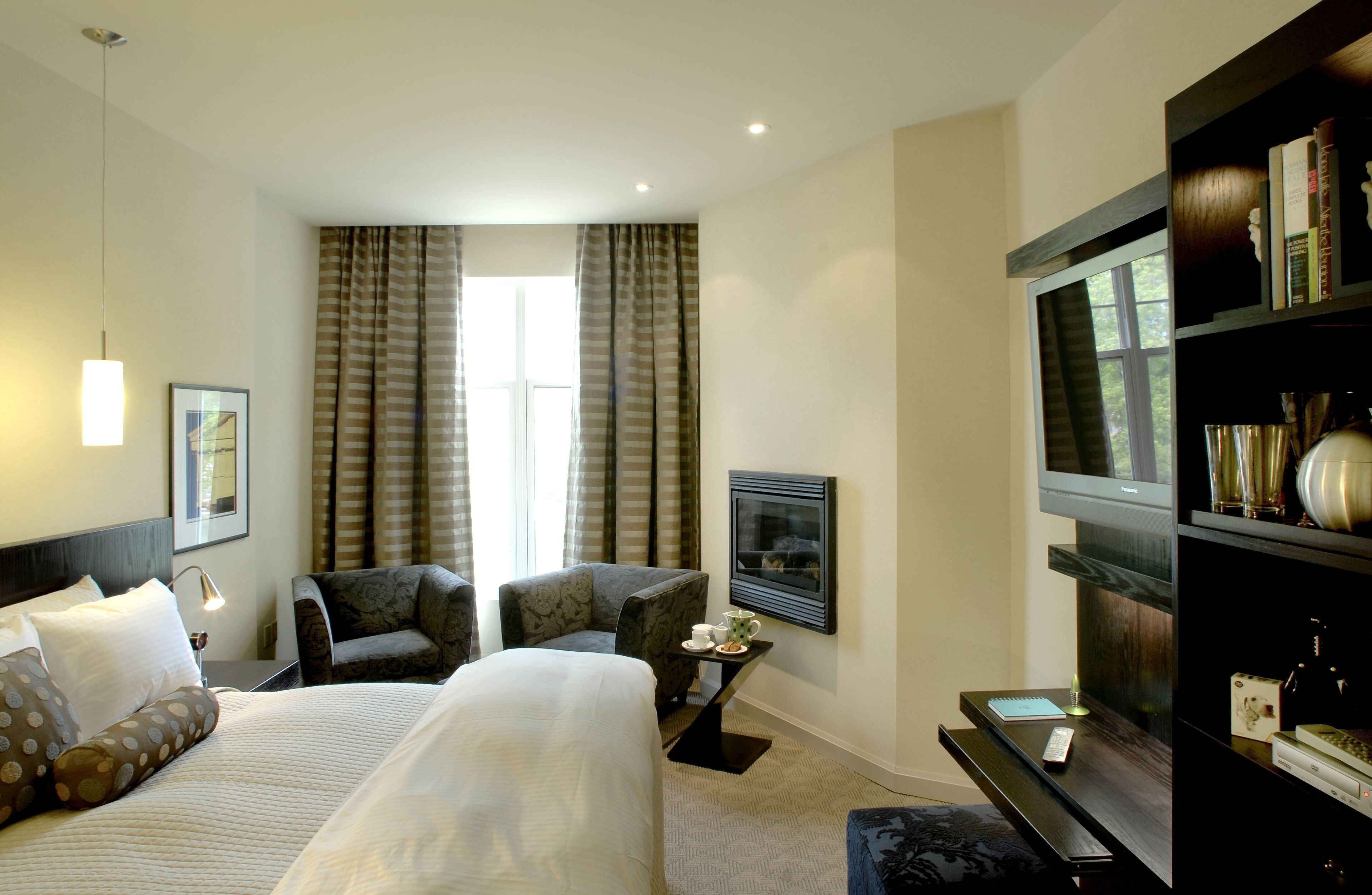 Una habitación de hotel con una cama grande, dos sillones, una mesita con lámpara, un televisor y una estantería.