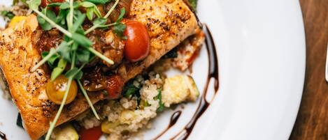Un plato de comida con un trozo de pollo a la parrilla cubierto de verduras y tomates cherry, acompañado de quinua y vegetales.