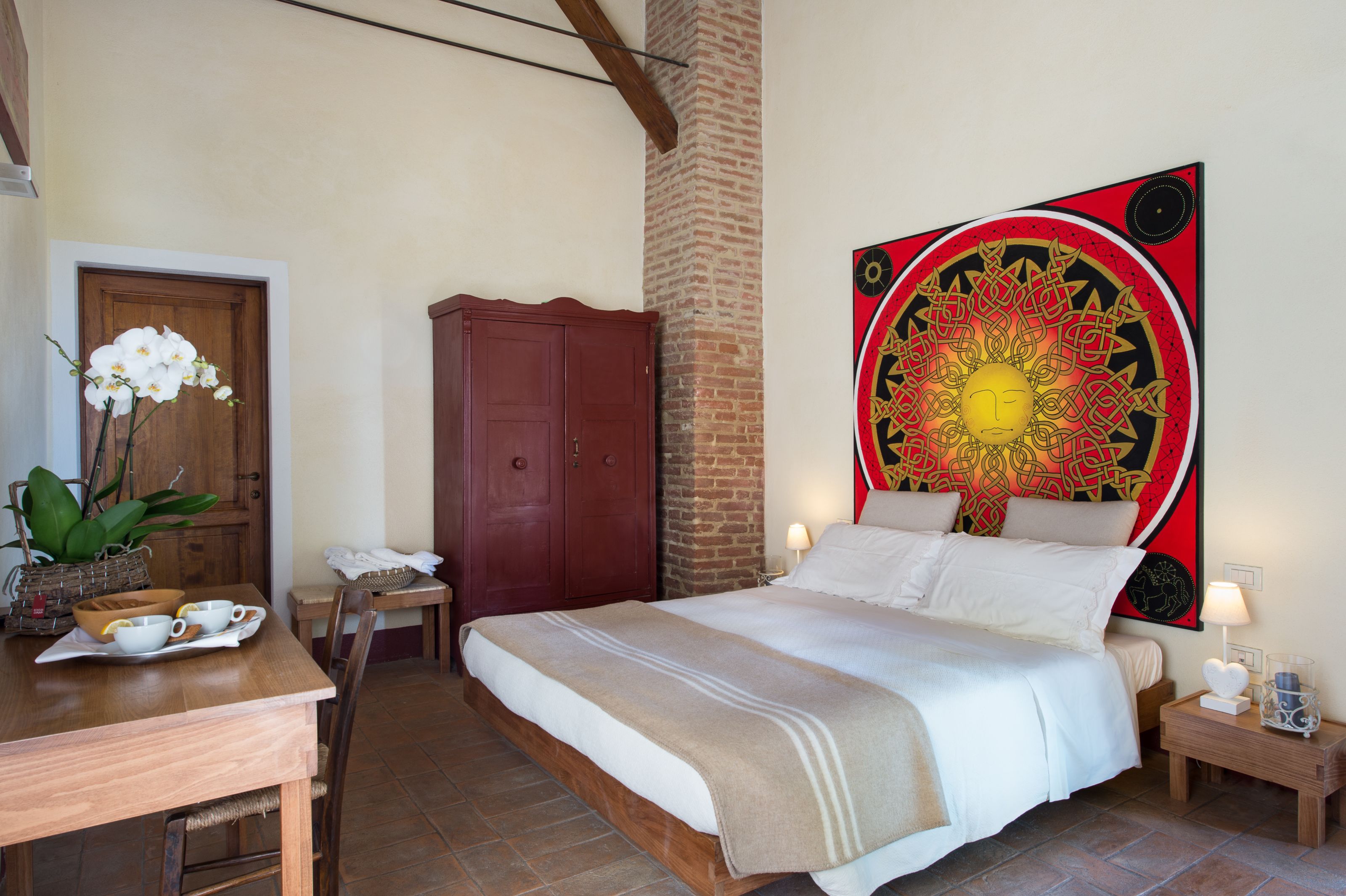A bedroom with a bed, a wooden desk, a chair, a dining table, and a large decorative wall piece.