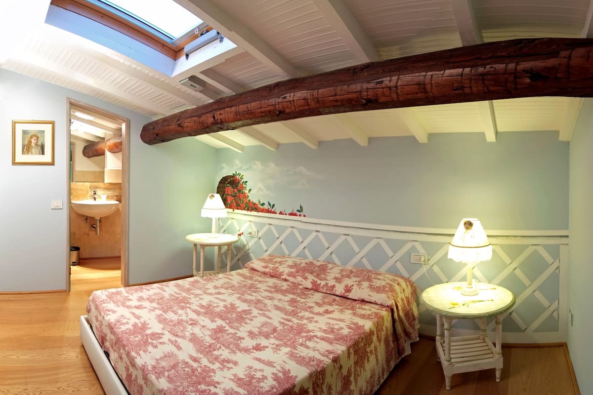 A bedroom with a skylight, wooden beams, a bed with floral bedding, two bedside tables with lamps, and a wall-mounted shelf with decorative items.