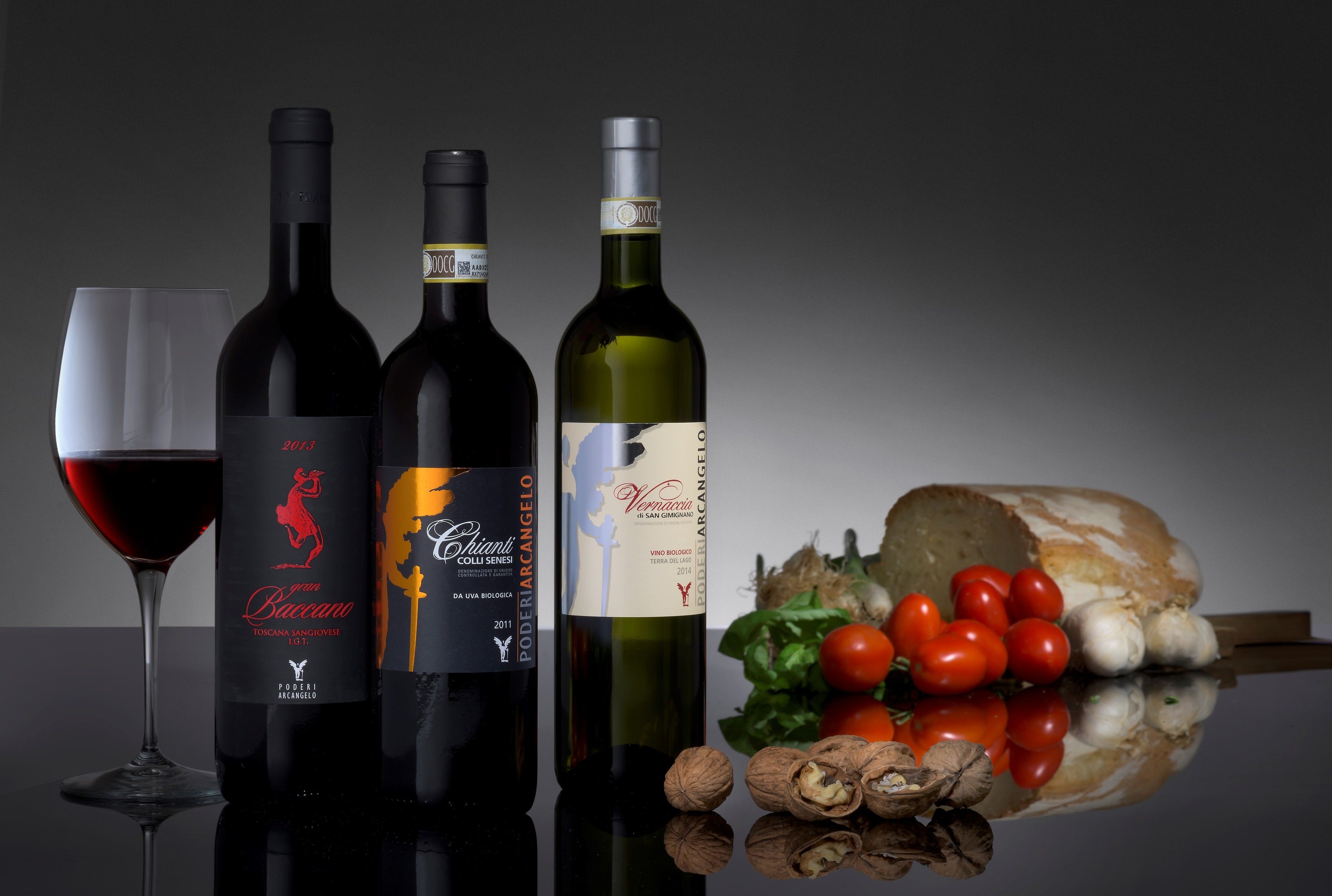 Tres botellas de vino y una copa de vino tinto sobre una superficie reflectante, con tomates, ajo y nueces.
