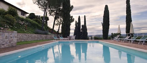 Ein Swimmingpool mit Liegestühlen und Sonnenschirmen, umgeben von Steinmauern und hohen Bäumen.