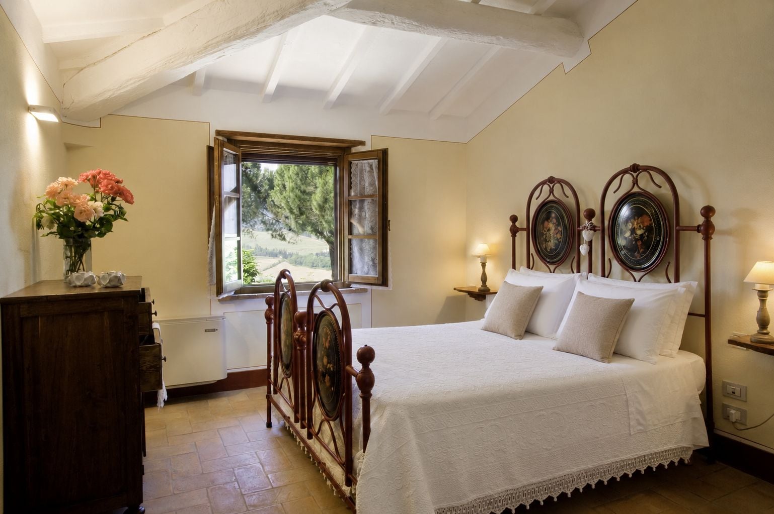 A bedroom with a wooden bed, a nightstand with a lamp, a dresser, and a window with a view of greenery.