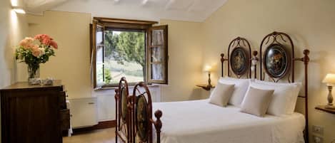 A bedroom with a wooden bed, a nightstand with a lamp, a dresser, and a window with a view of greenery.