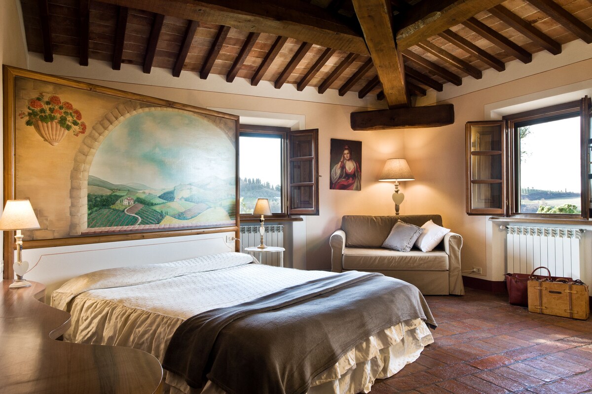 Un dormitorio con una cama grande, una silla, una mesa, una lámpara, un cuadro y vistas al exterior.