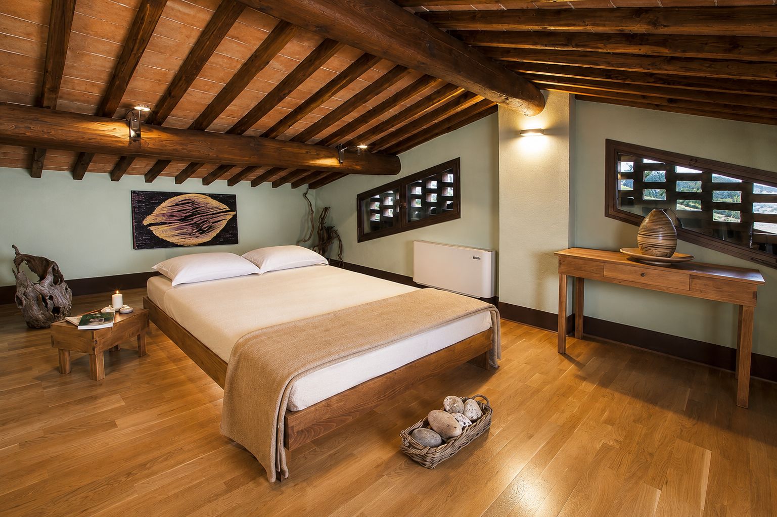 Un dormitorio con techo de madera, una cama con manta color beige, una mesita de noche con un libro, una repisa en la pared con objetos decorativos y una ventana con vistas.