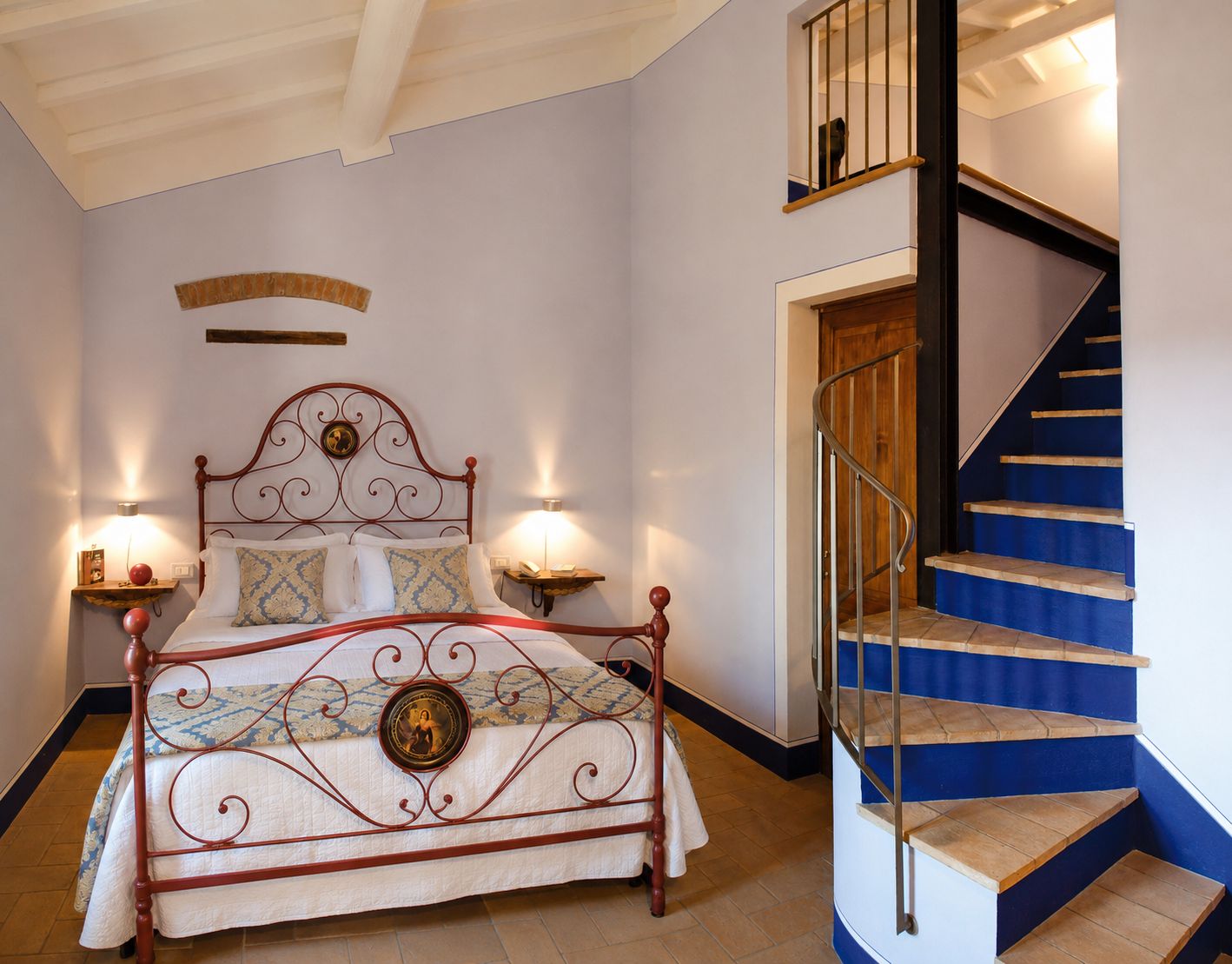 A bedroom with a metal-framed bed, two bedside lamps, a wooden headboard, and a blue staircase with a wooden railing.