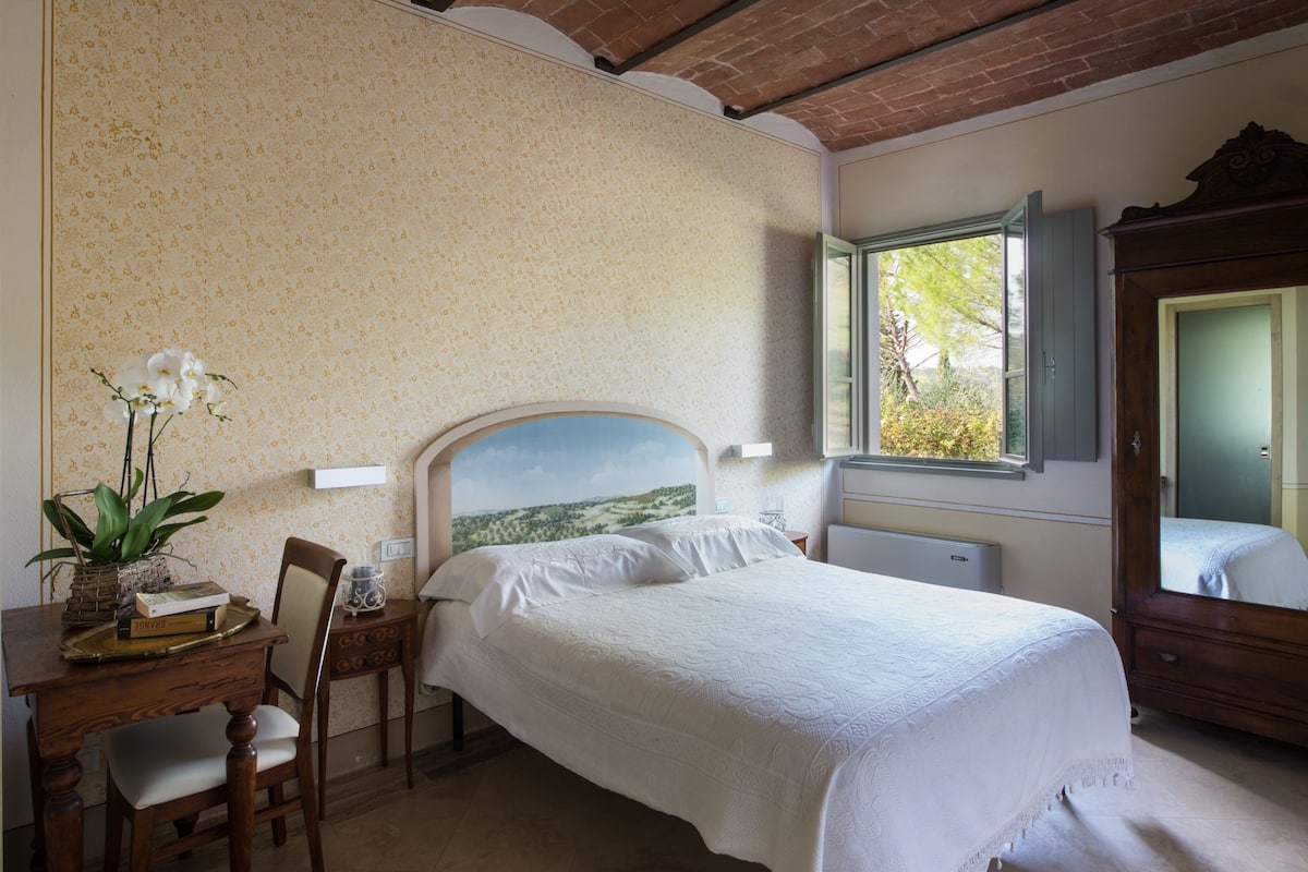 A bedroom with a bed, a desk with a chair, a dresser, and a window with a view of greenery.