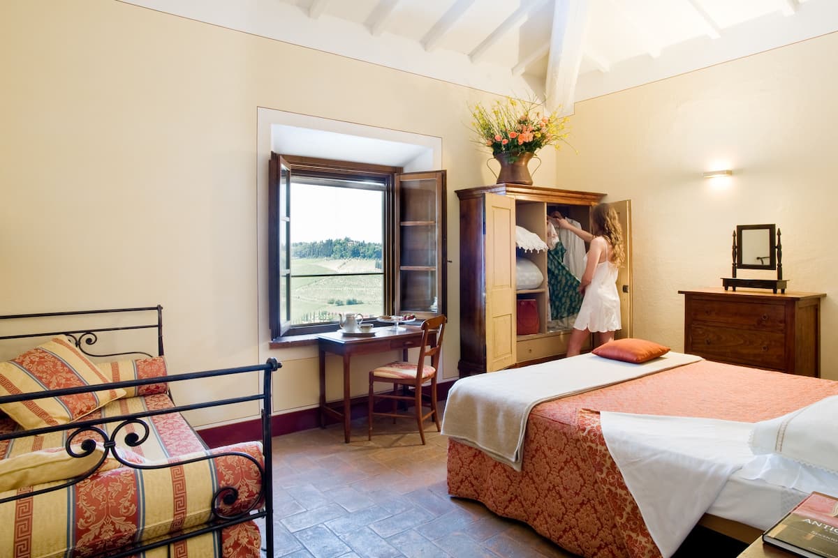 A bedroom with a bed, a desk, and a chair. There is a window with a view of a field.