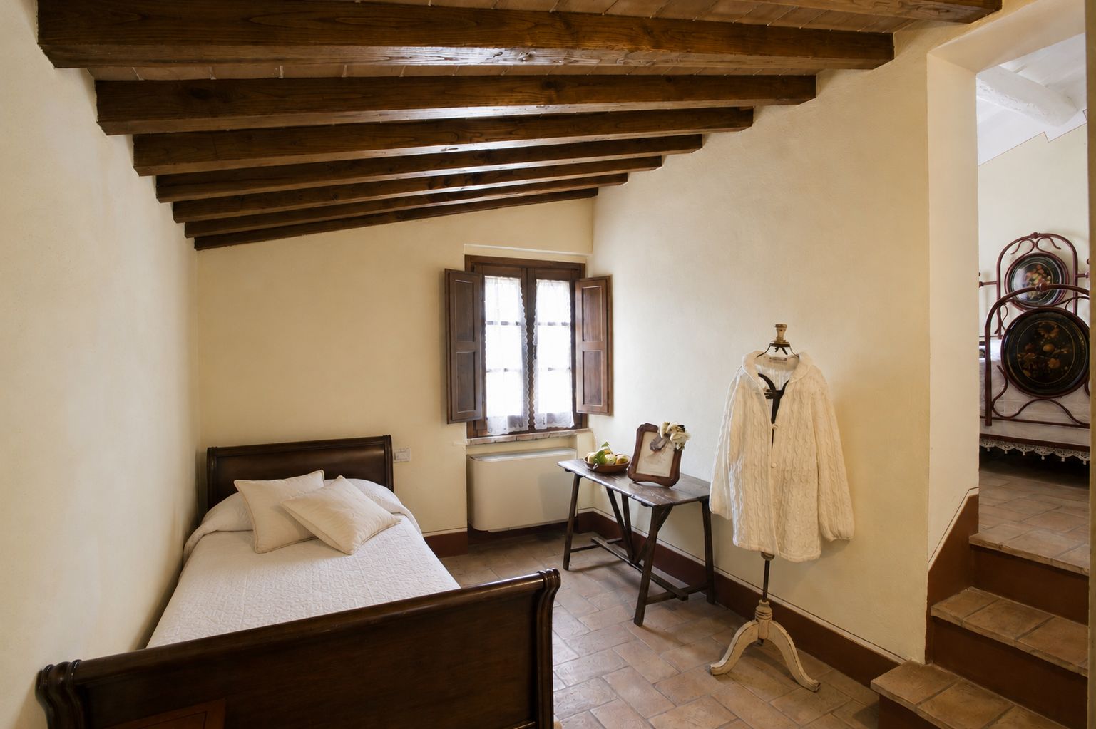 Un dormitorio con cama de madera, una mesita con un marco de foto, un abrigo en un perchero y una ventana con contraventanas.