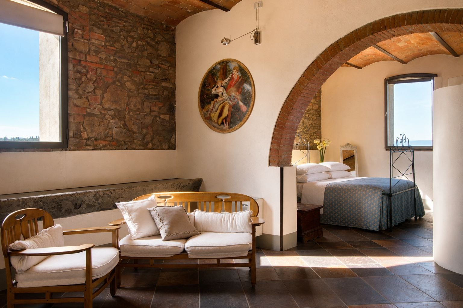 A room with a stone wall, a bed with a patterned bedspread, a wooden bench, and a painting on the wall.