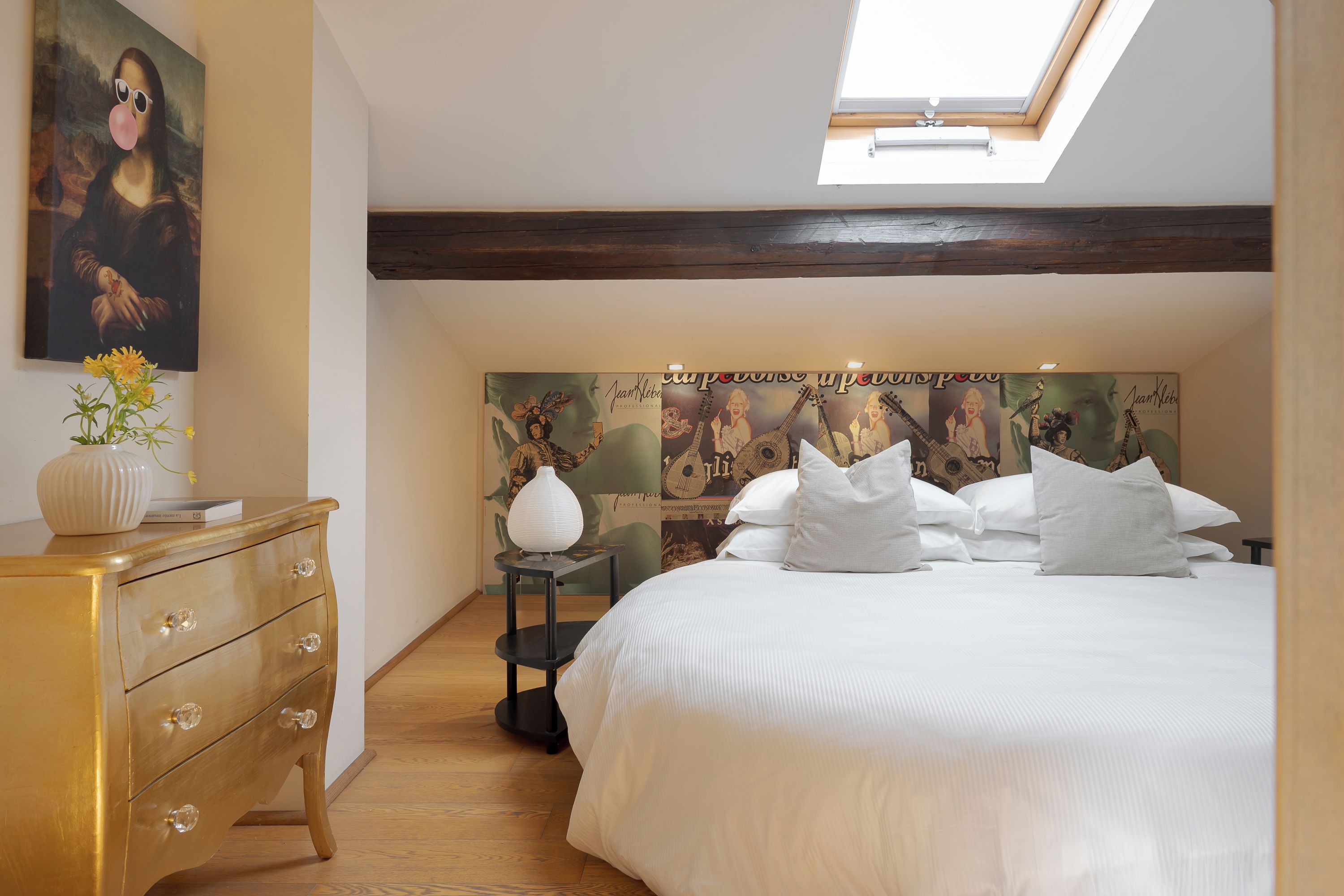 A bedroom with a bed, a dresser, a vase with flowers, a skylight, and a wall with a poster.