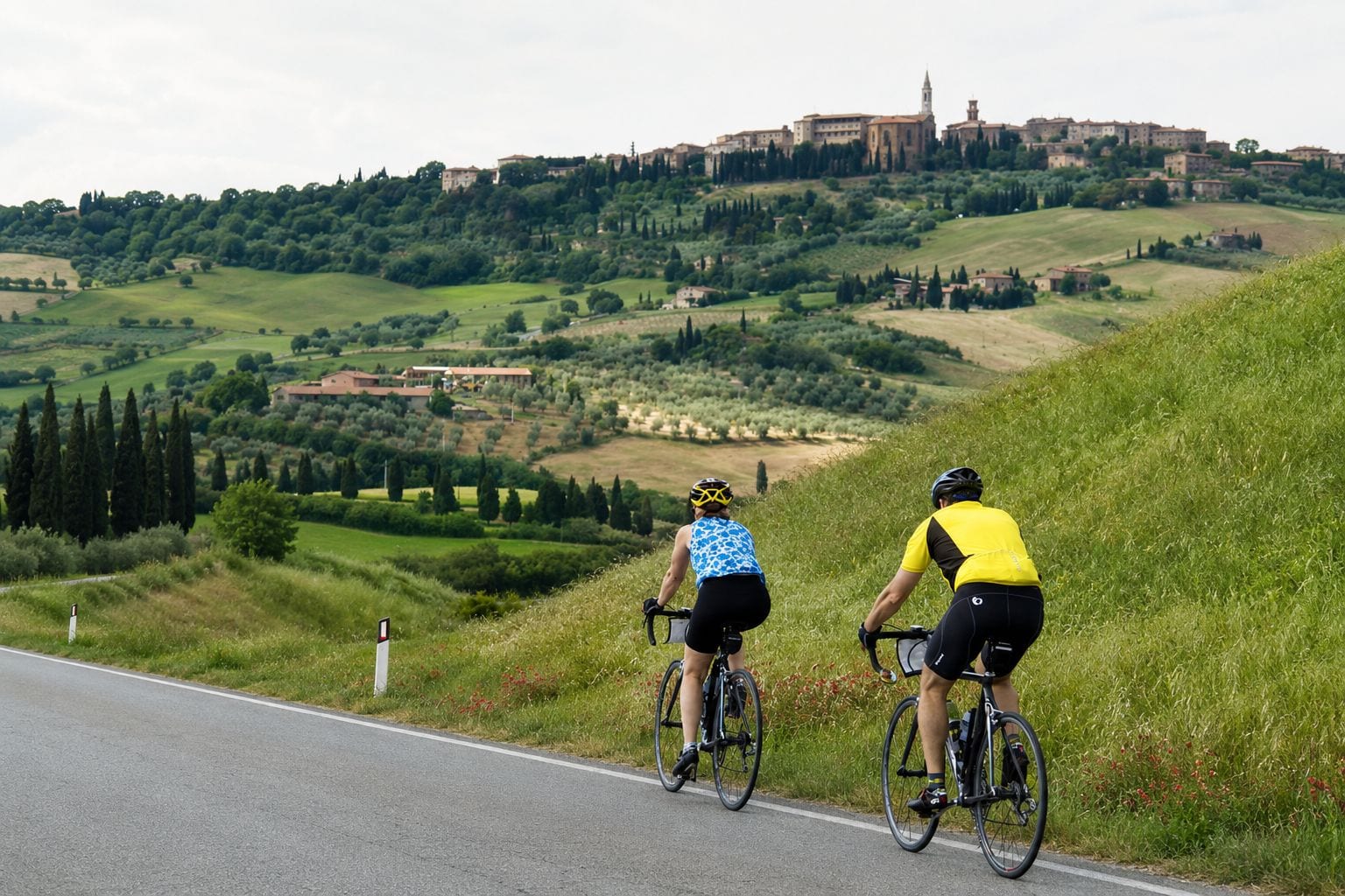 Two cyclists on a road with a view of a hilltop town and castle in the background.